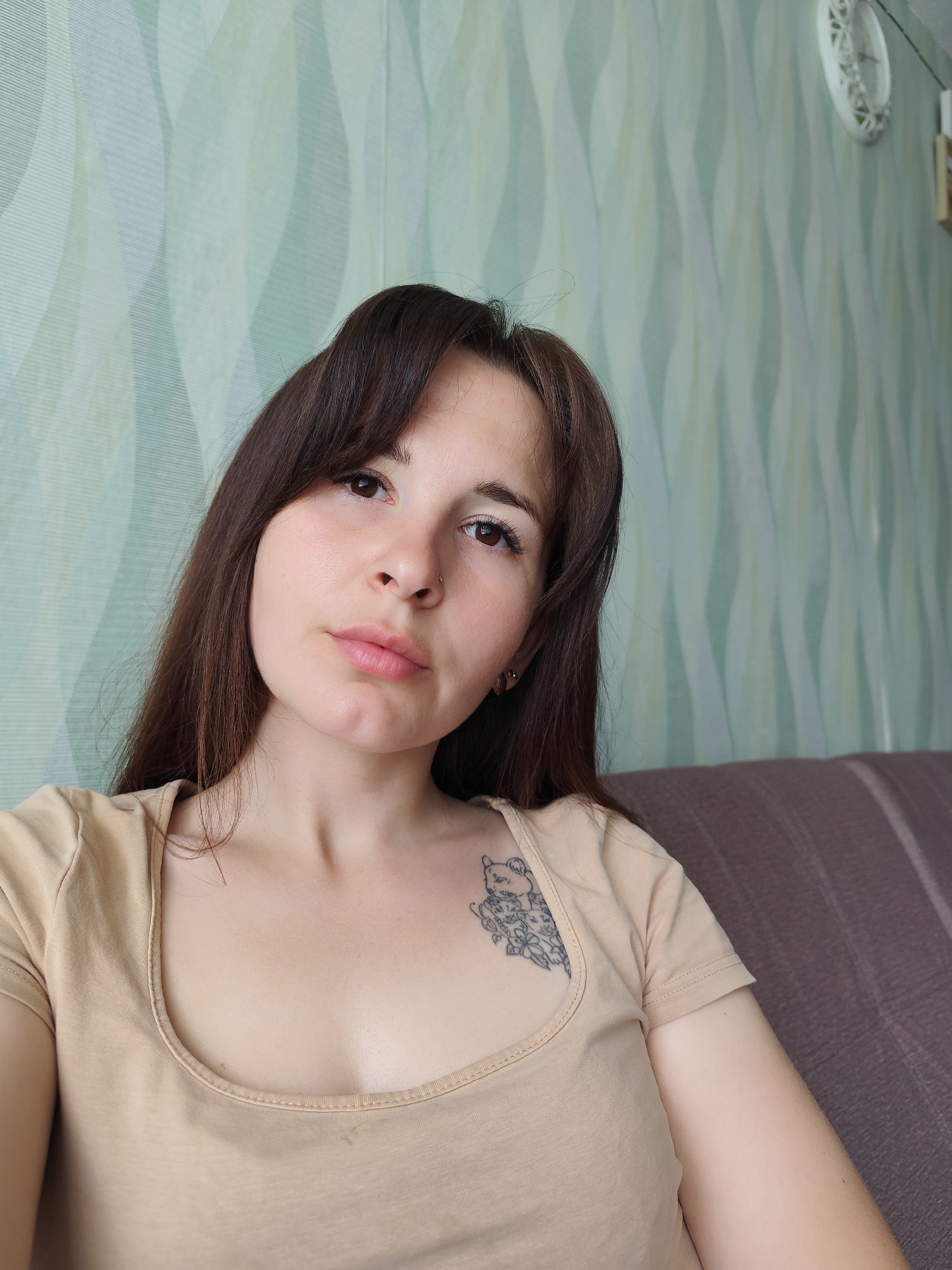 A woman is in a room with wallpaper. The wallpaper has an aqua and white vertical wavy pattern. The woman has long dark hair with bangs, fair skin, brown eyes, and a nose ring. She is wearing a light beige t-shirt. A small tattoo of a cat with flowers is visible on her upper chest. The room also contains a clock on the wall. A portion of a brown couch is visible. The city of Фоки, Russia, cannot be verified from the image.