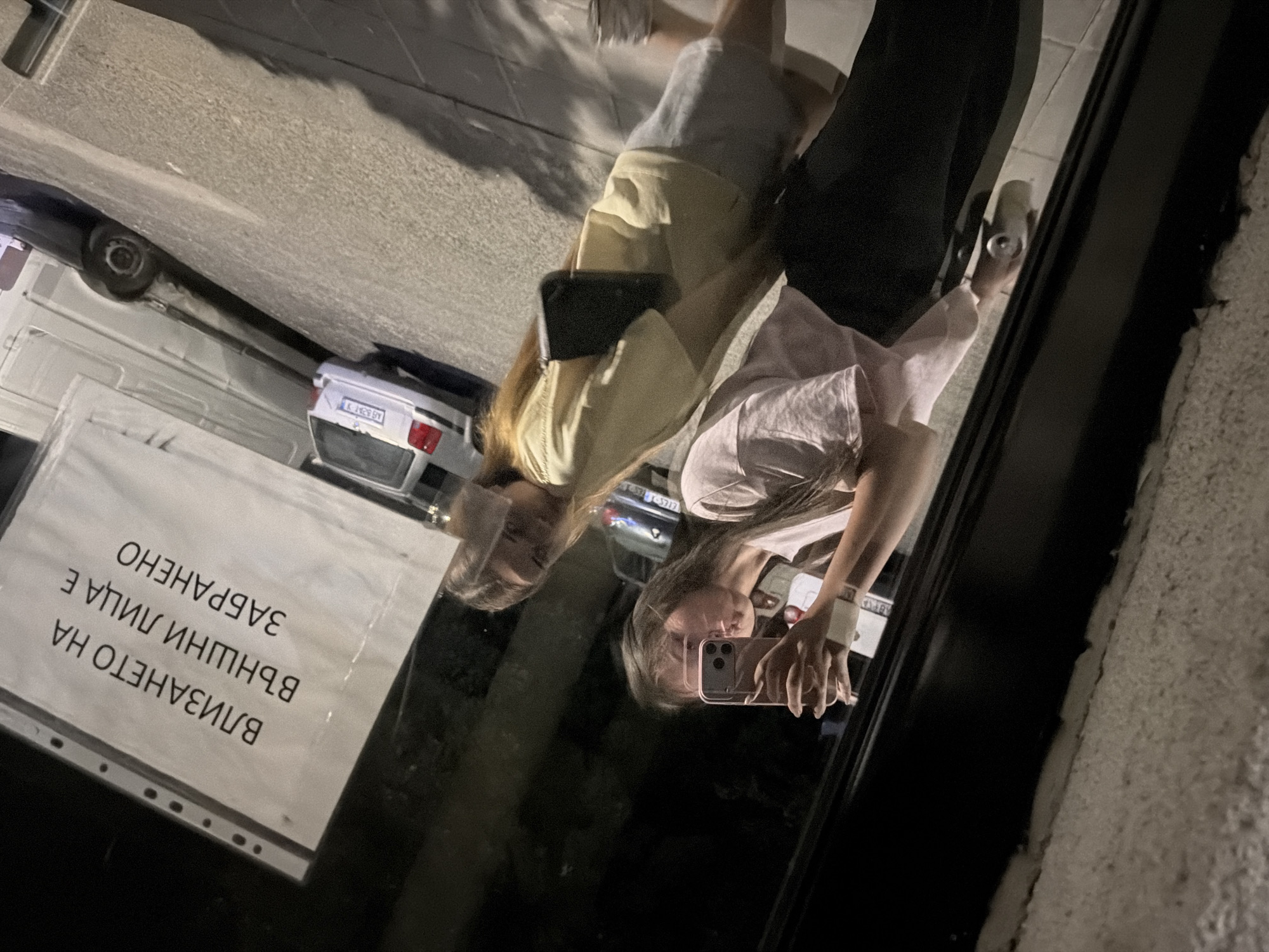 The image shows two young women taking a selfie in front of a reflection. The woman on the left is holding a phone and wearing a light pink long-sleeved shirt and dark pants. The woman on the right is wearing a light yellow t-shirt and grey shorts, and is holding a black clutch. In the background, a white van and a silver car are visible, along with a sign in Bulgarian that reads "Entrance of external persons is forbidden." The scene appears to be outdoors at night, possibly in a parking lot or street in Novi Pazar, Bulgaria.