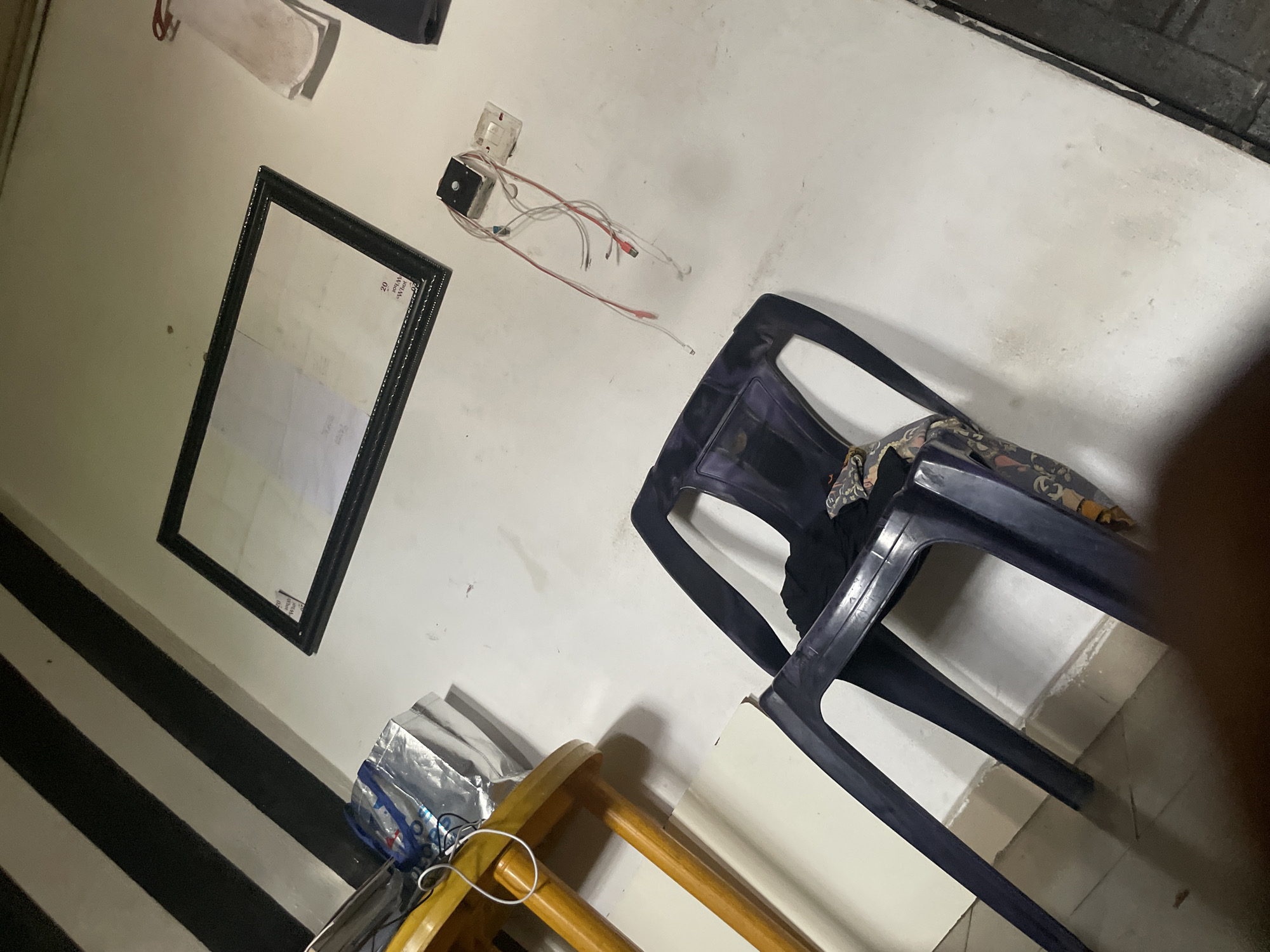 The image depicts a dimly lit room, likely in Ihiagwa, Nigeria, characterized by a white wall with black and white striped diagonal markings near the ceiling. A black framed picture hangs on the wall. To the left, a yellow wooden table with a white sheet and a silver bag is visible. In the foreground, a dark plastic chair is present. A person's leg is partially visible in the bottom right corner, suggesting the photographer's presence. The scene appears to be a residential or storage area, with a slightly cluttered and unkempt appearance.