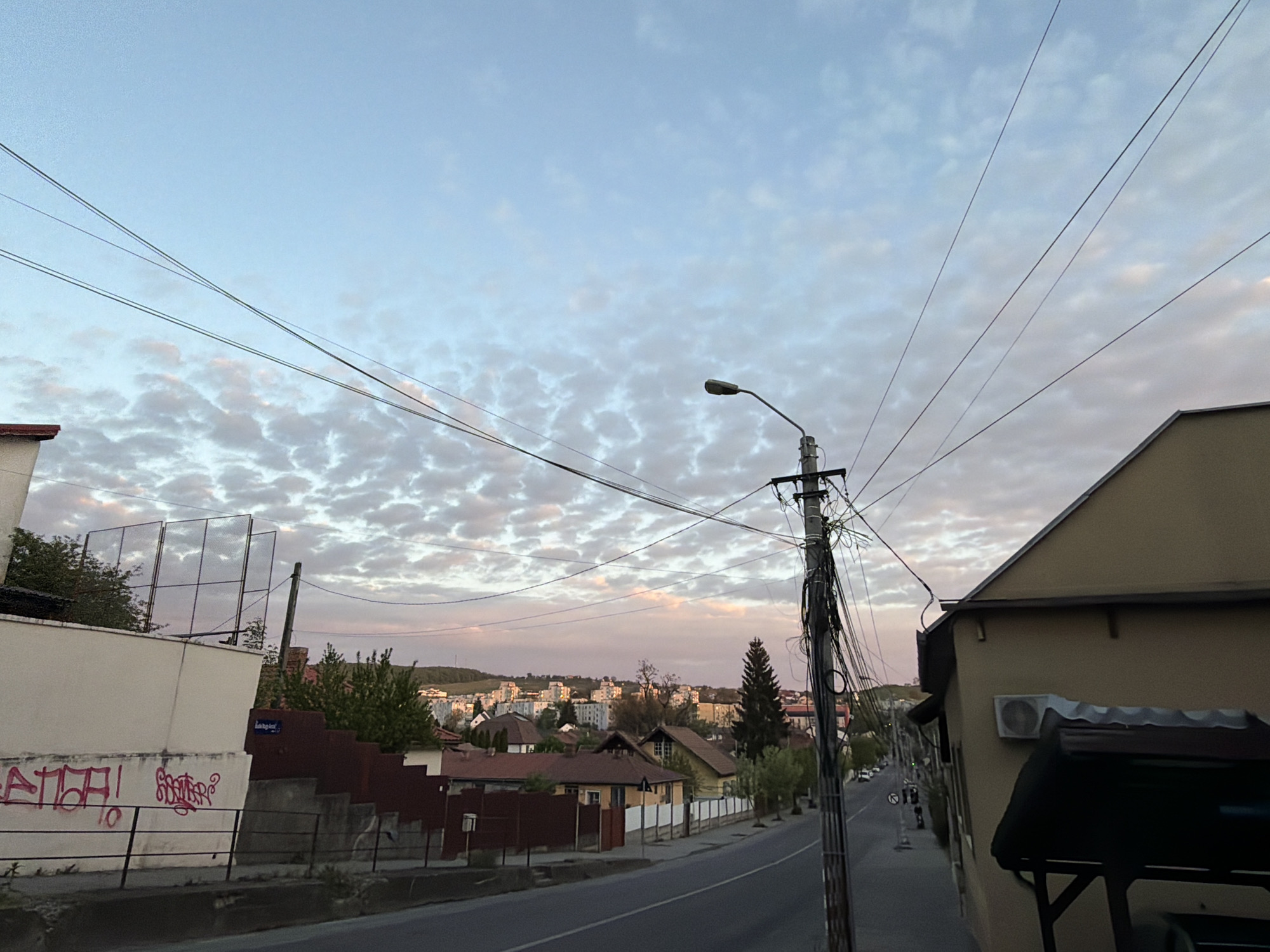 The image presents a street scene in Târgu Mureș, Romania. The sky is a mixture of blue and altocumulus clouds, suggesting either dawn or dusk. Electrical wires crisscross above the street.
On the left is a white building with graffiti on its wall and a rusty metal fence in front. There is a sign in Romanian with white lettering on a blue background, near a set of stairs. On the right is a building with a tan facade.
A utility pole with a streetlight is centrally located, with numerous wires attached. The street runs between buildings, with houses visible on the other side and in the distance. In the distance, there are several mid-rise apartment buildings. A few trees are visible among the houses.
On the street there are a couple of individuals walking, but it's difficult to distinguish their features. Several parked cars are visible in the distance on the street. The photograph is taken at eye level.