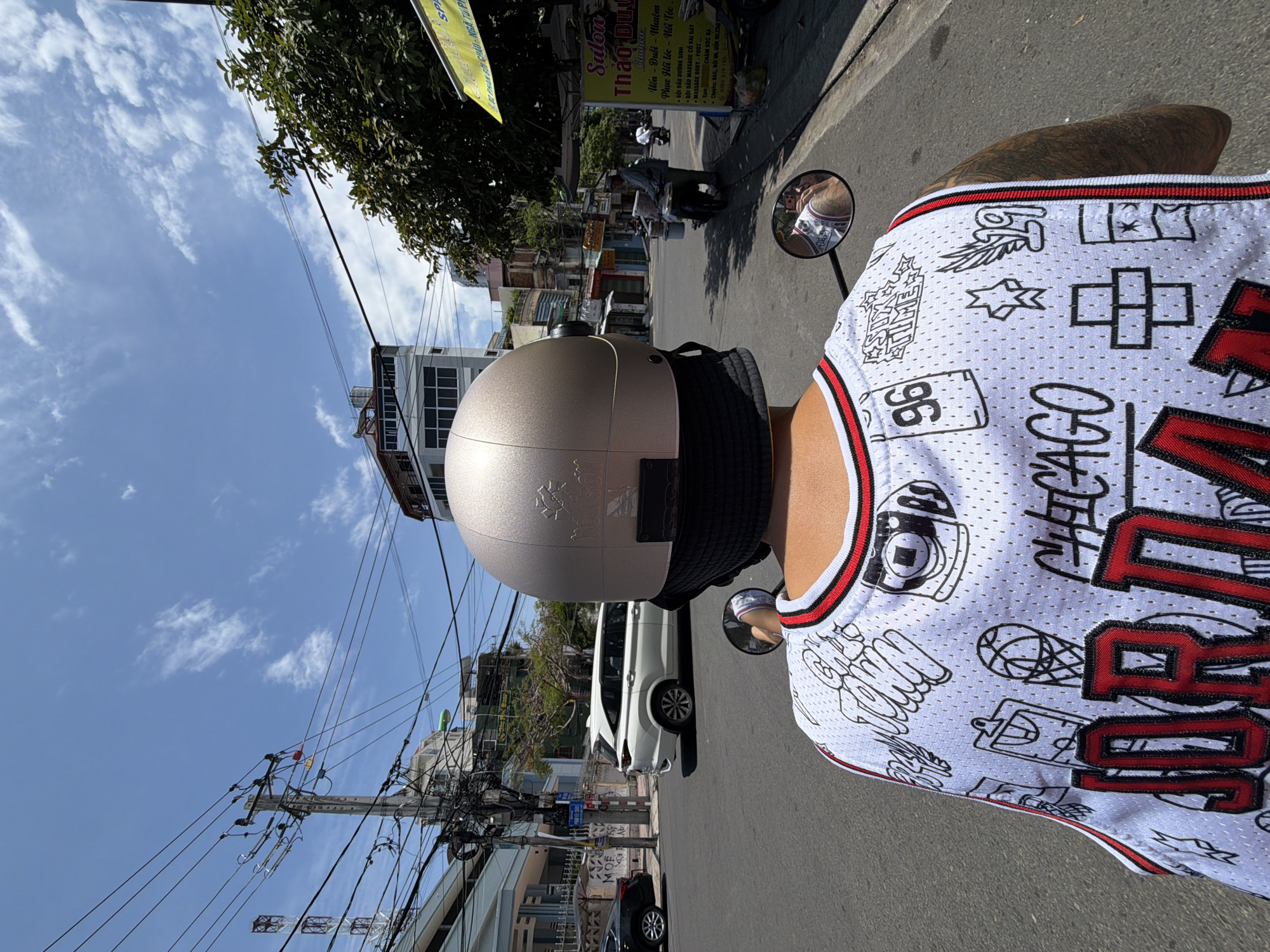 The image is taken from the perspective of someone riding a motorbike in Nha Trang, Vietnam. The rider is wearing a white basketball jersey with "JORDAN" prominently displayed in red lettering and various graphic designs. They are also wearing a gold helmet. In the background, there are typical city elements including buildings, power lines, a white SUV, and other motorbikes. A yellow sign for "Salon Thảo Duy" is visible on the right side of the street. The sky is blue with scattered clouds.