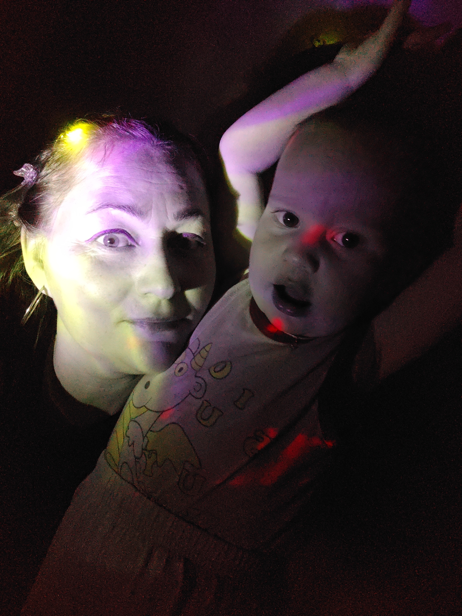 The image shows a woman and a baby. The woman has fair skin, dark hair, and is wearing hoop earrings. A yellow light is visible in her hair. The baby is wearing a light-colored outfit with cartoon graphics on it. Red light is illuminating the baby's face and neck. The background is dark and grainy. The location is not evident from the image.