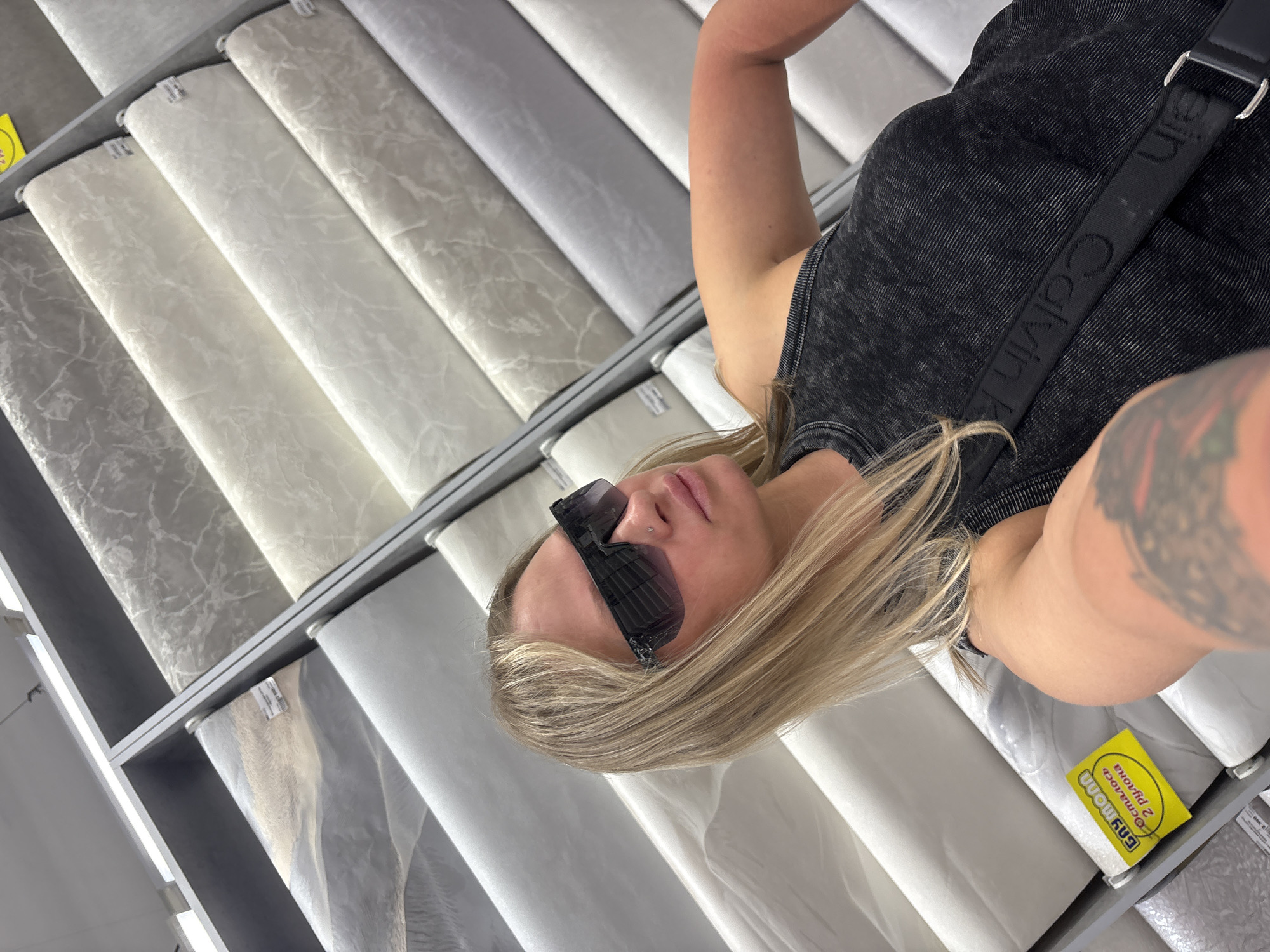 A person with blonde hair, wearing sunglasses and a black tank top with a "Calvin Klein" strap, is in a shop looking at a wall-mounted display of rolls of wallpaper, with some rolls having yellow labels with Russian text. The location is most likely a home improvement or wallpaper store in the city of Perm, Russia.