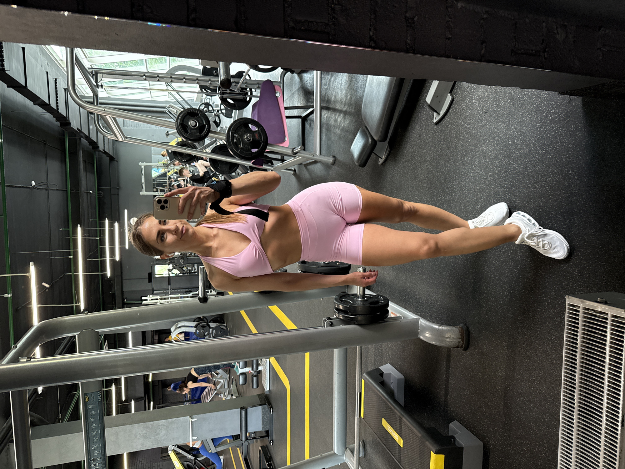 The image depicts a woman in athletic wear taking a selfie in a gym. She is wearing a pink sports bra and pink shorts, along with white sneakers. She has on black wrist wraps. The gym environment is visible in the background, with various weight machines, benches, and exercise equipment. The floor is black rubber matting, and there are yellow markings on the floor. The overall impression is one of a person engaged in fitness activities at a gym. The location is identified as Saint Petersburg, Russia.
