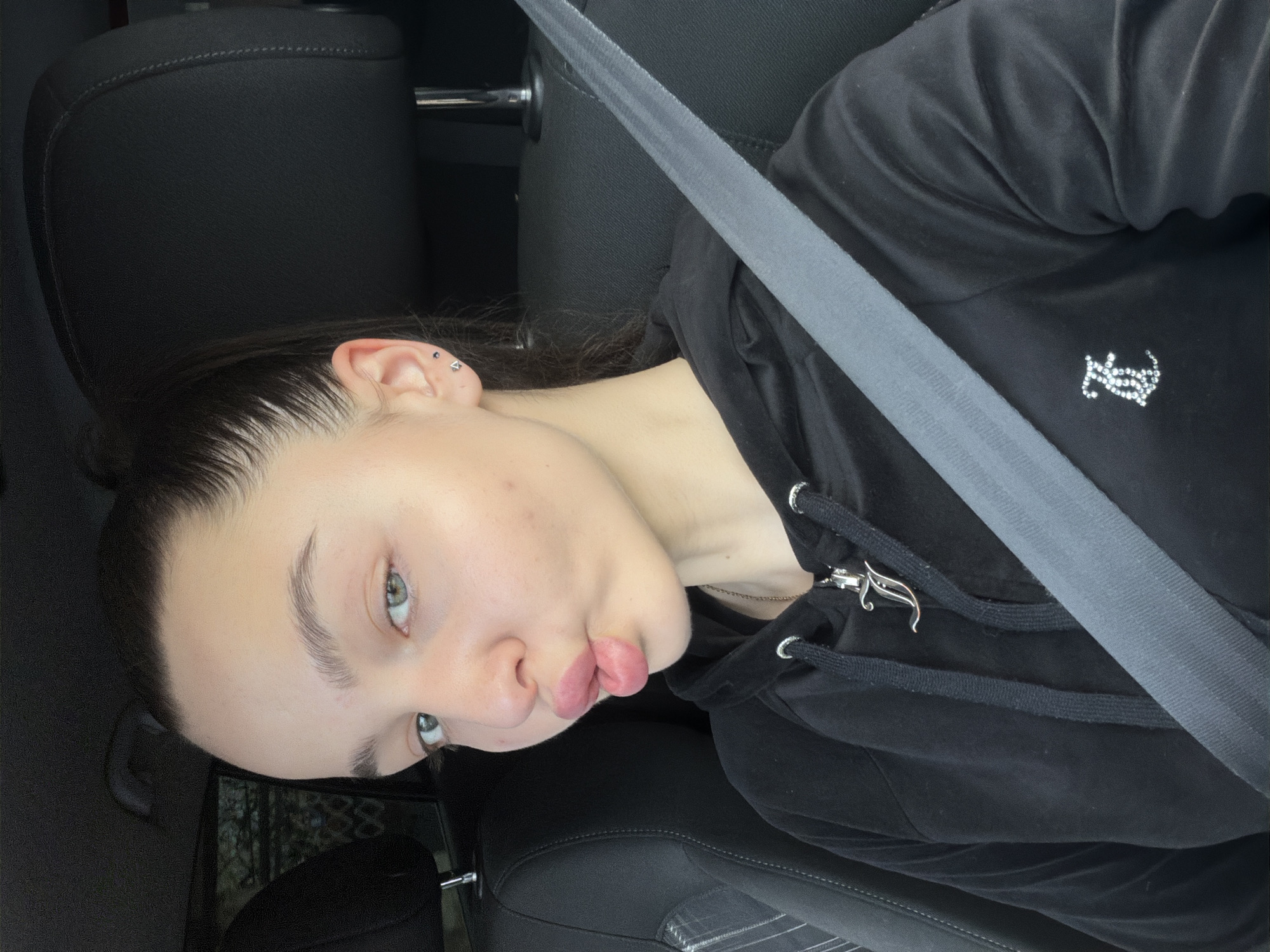A young woman is in a car, wearing a black hoodie with a bedazzled logo. She is wearing a seatbelt and pouting. Two piercings are visible in her ear. The location is likely not Busmantsi, Bulgaria as there is no evidence to support that claim.