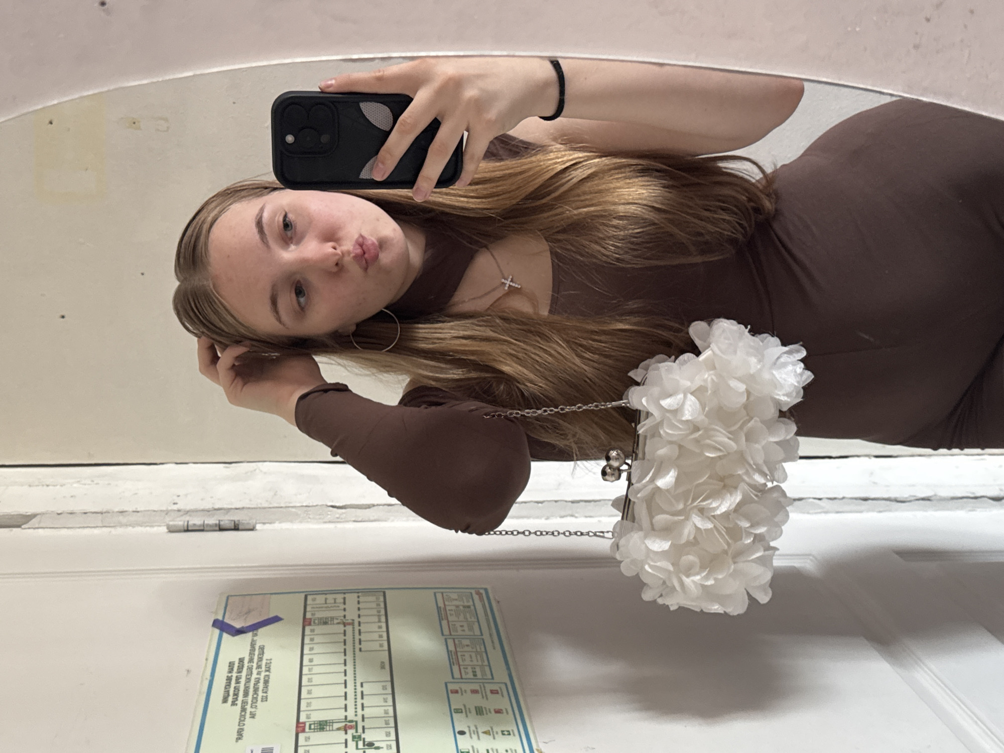 The image shows a young woman in a brown one-piece garment, holding a white floral clutch purse and a smartphone. She is looking at her reflection in a mirror and has her hand on her hair. She is wearing a cross necklace and hoop earrings. Behind her, a door with a poster is visible, along with part of a white door frame. The overall setting appears to be an indoor space, possibly a bathroom or hallway, with a wall that shows signs of wear. The poster on the wall is an evacuation plan, indicated by symbols and text, common in public buildings. The image does not provide enough information to determine the specific location within the city of Perm, Russia.