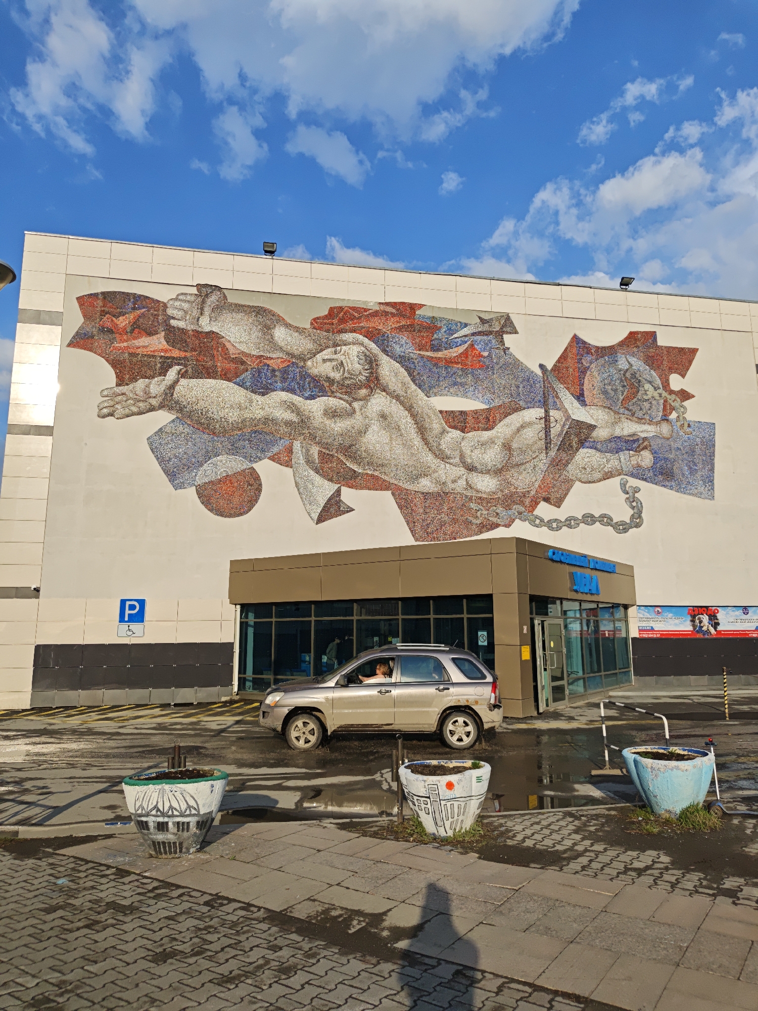 The image shows the exterior of a building in Yekaterinburg, Russia. The building has a large mosaic mural on its facade depicting a nude male figure and a metal anchor shackled with chains. A car is positioned in front of the building entrance. In the foreground, there are three large, decorated planters. The weather appears to be sunny with blue skies and scattered clouds. The ground is wet, reflecting light.