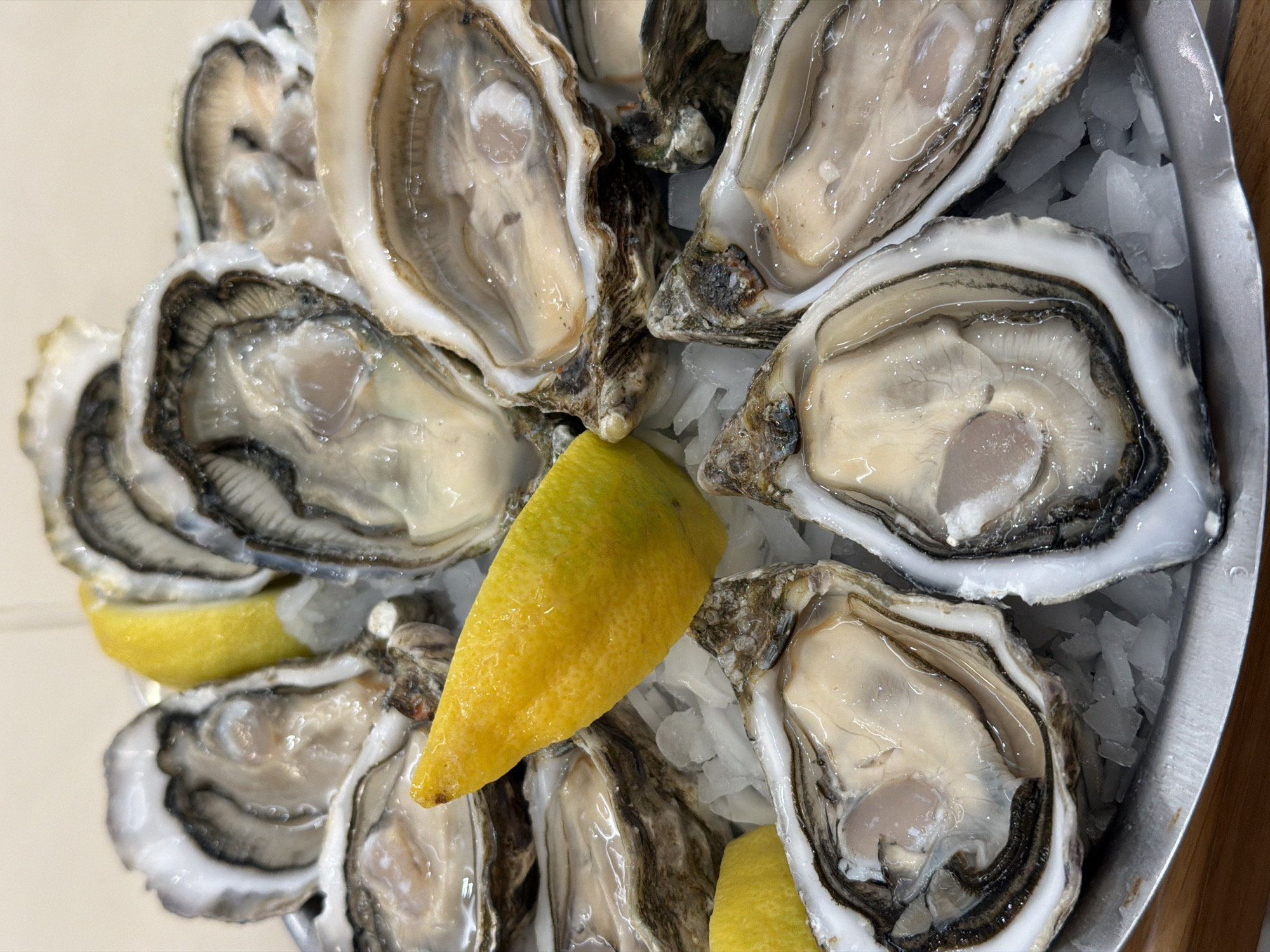 The image shows a platter of fresh oysters on ice, garnished with lemon wedges. The oysters are open, revealing their plump, gray-beige bodies within their shells. The shells are varied in color and texture, ranging from gray to brown with intricate patterns. Small shards of ice fill the spaces between the oysters, and a few bright yellow lemon wedges are interspersed among them. The platter appears to be made of silver or stainless steel. The image composition and subject matter suggest the scene is likely at a restaurant, possibly in Paris, France, given the association of oysters with French cuisine.