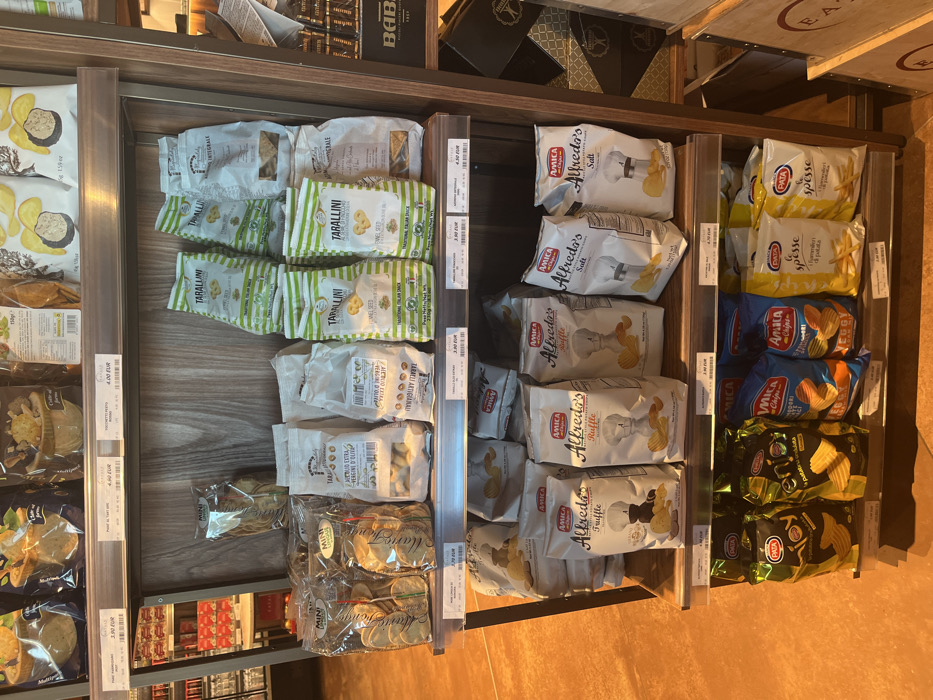 The image displays an indoor scene, likely within a grocery store or specialty food shop in Dorno, Italy, given the context. The primary subjects are various packaged food items, predominantly snacks and crackers, arranged on brown wooden shelves with clear plastic dividers.
On the left side of the shelving unit, there are bags of "TARALLINI" in green and white packaging, some labeled "WHOLEGRAIN" or "ONE SEED." Below these are white bags of "TARALLI ARTIGIANALI," including a variety labeled "EXTRA VERGINE OLIVA." Further down and to the left, there are clear packages of "MINI" branded crackers or snacks. Adjacent to these, on the far left, are larger white and yellow bags of potato chips, possibly featuring truffle or other flavors, some with prices like "4.00 EUR" or "3.90 EUR".
The right side of the display features several brands of potato chips. The top shelves hold white bags of "AMICA Alfredo's Salt" chips, recognizable by the salt shaker graphic. Below these are "AMICA Alfredo's Ruffle" and "AMICA Alfredo's Truffle" chips, distinguished by their respective flavor icons. Further to the right, yellow bags of "PATA spesse" chips are visible, followed by blue bags of "AMICA Chipi" or "AMICA Chips ENERGY." The bottom right shelves contain dark green/black bags of "PATA" brand chips.
Many products have visible price tags, such as "3.90 EUR," "3.00 EUR," "2.80 EUR," and "2.70 EUR," indicating that items are for sale. In the background, behind the shelves, there are dark wooden elements and some cardboard boxes, one with the partial text "BAB" and another "EA." The lighting suggests an indoor setting, and no people or specific activity are visible, only the neatly organized merchandise. The prominent Italian text on many packages further reinforces the location context.