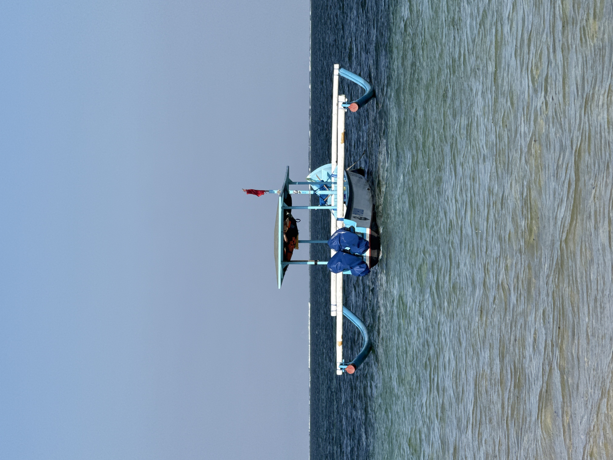 A traditional Indonesian outrigger boat with a blue canopy and a red flag is moored in the clear, shallow waters of Denpasar, Bali. The boat features two blue cushioned seats and two blue float bags. The calm sea reflects the clear blue sky. The boat is the central subject of the image.