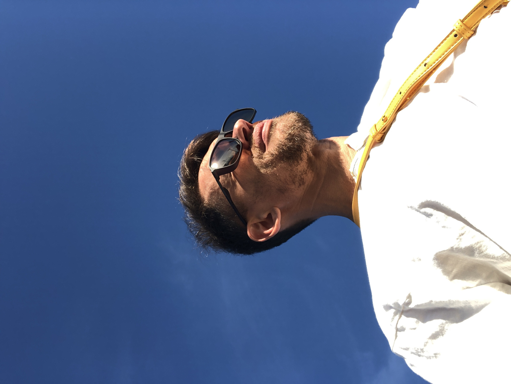 A low-angle, medium shot captures a man from the chest up against a bright blue sky. The man, positioned off-center to the left, wears dark-framed sunglasses and has a short, brown beard and dark hair. He is looking upwards and to the right, with his mouth slightly open and a faint smile visible. A thin, yellow strap is visible diagonally across his white, loose-fitting shirt, which appears to be made of a light, possibly linen or cotton, material. The sunlight strongly illuminates the man's face and shirt, creating bright highlights and some shadowed areas. The sky is clear and a deep, vibrant blue, with no clouds visible. The overall impression is of a sunny day outdoors.