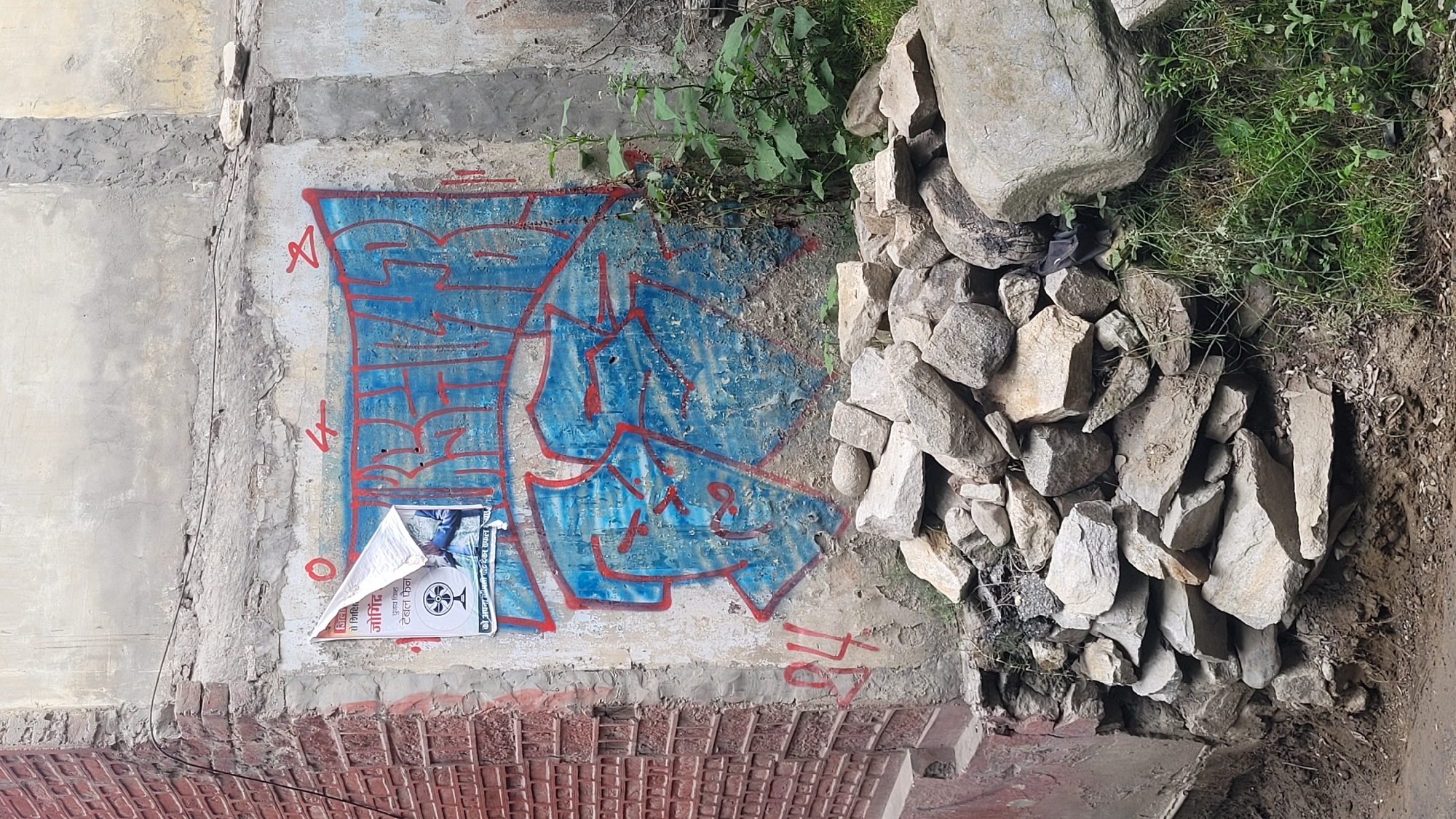 The image depicts a weathered concrete wall in Vashisht, India, with graffiti and a torn poster. The graffiti, predominantly blue with red outlines, covers a significant portion of the wall. It appears to be a stylized lettering or tag. A partially ripped poster, featuring an image of a fan and text in a non-Latin script, is affixed to the wall near the graffiti. To the left of the wall is a section of red brickwork with a patterned texture. In front of the wall, a pile of rocks and debris has accumulated, with some vegetation growing around and through it. The ground in the foreground is dirt. The overall scene suggests an urban environment, possibly an alleyway or a neglected exterior wall.