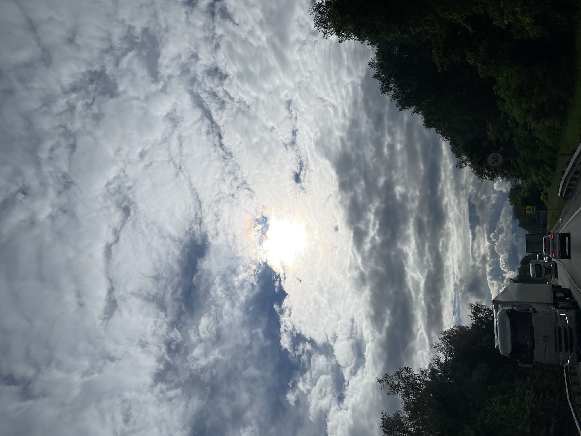 The image shows a view looking upwards and slightly to the right, capturing a sky filled with textured, layered clouds, partially obscuring the sun. The scene includes a road with vehicles, including a white truck, and trees lining the roadside. A road sign with a '50' speed limit is visible. This image was not taken in the city of Ol'khovka, Russia.