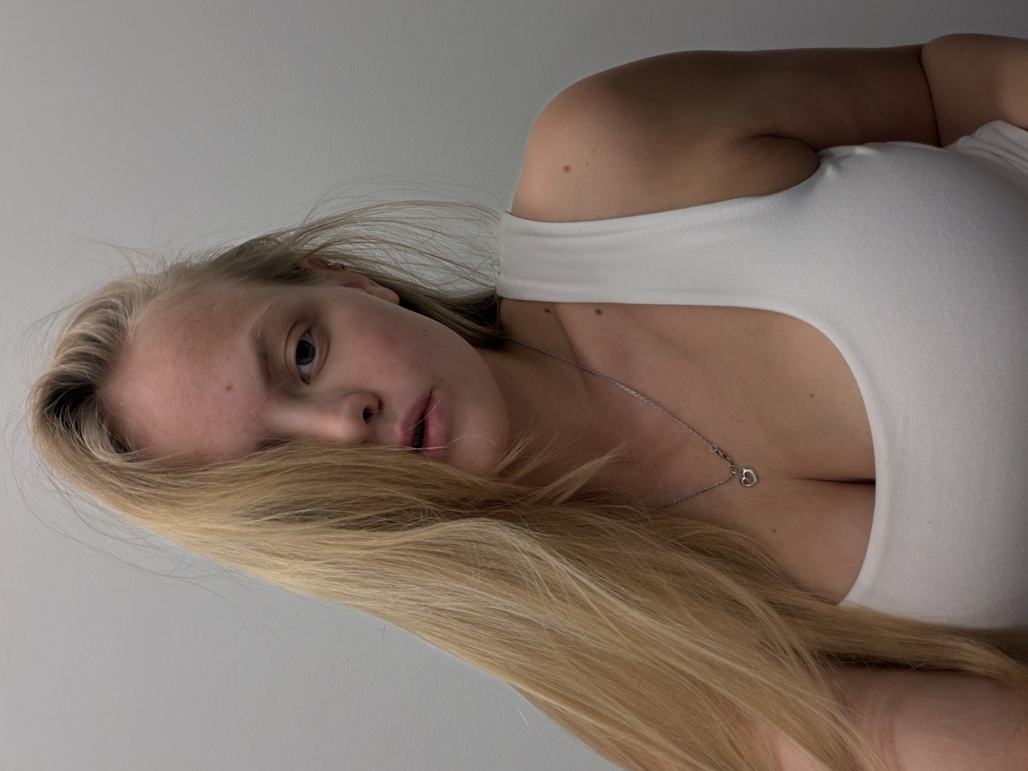 A close-up portrait depicts a young person with long, straight blonde hair, positioned as if lying down and gazing directly towards the viewer. The image is rotated 90 degrees counter-clockwise, with the subject's head on the left and torso extending to the right. The person is wearing a white, form-fitting tank top or camisole that reveals some cleavage. A delicate silver chain necklace with a small heart-shaped pendant rests against the skin.
Long blonde hair cascades across the lower part of the frame, partially obscuring the face, though one eye and slightly parted lips are clearly visible. The background is a plain, light grey or off-white surface, indicating an indoor setting. The lighting is soft and even, highlighting the subject's features and the texture of their hair. The person's expression appears neutral or slightly pensive. There are no visible location cues within the image itself to suggest the given context of Izhevsk, Russia, nor any discernible text. No specific activity beyond posing for the photograph is evident.