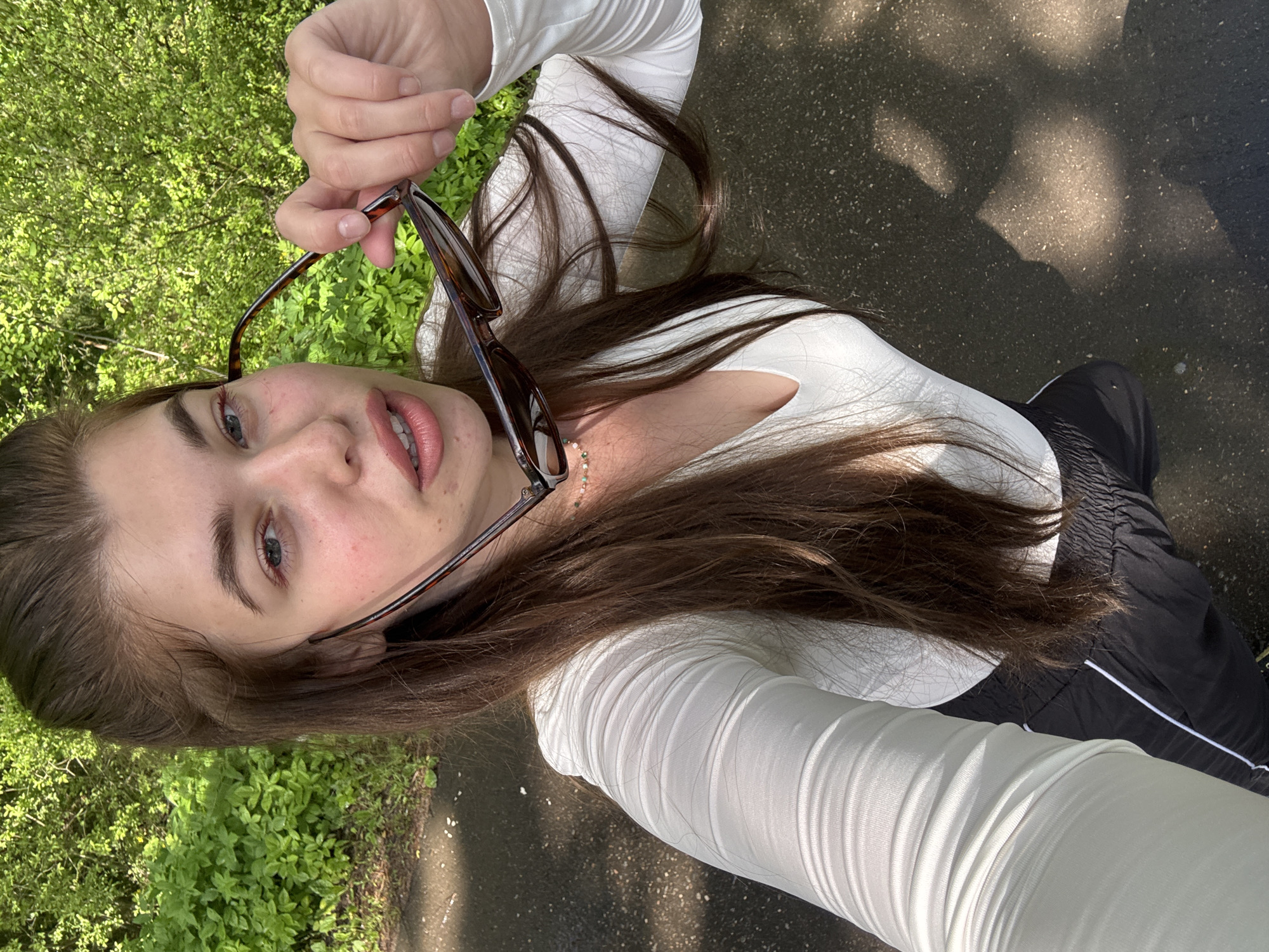 A young woman lies on a paved surface in Шевлягино, Russia, near green foliage. She holds a pair of brown-rimmed eyeglasses in her right hand. She wears a white, long-sleeved top and black athletic pants with a white stripe. Her long, brown hair is spread around her head.