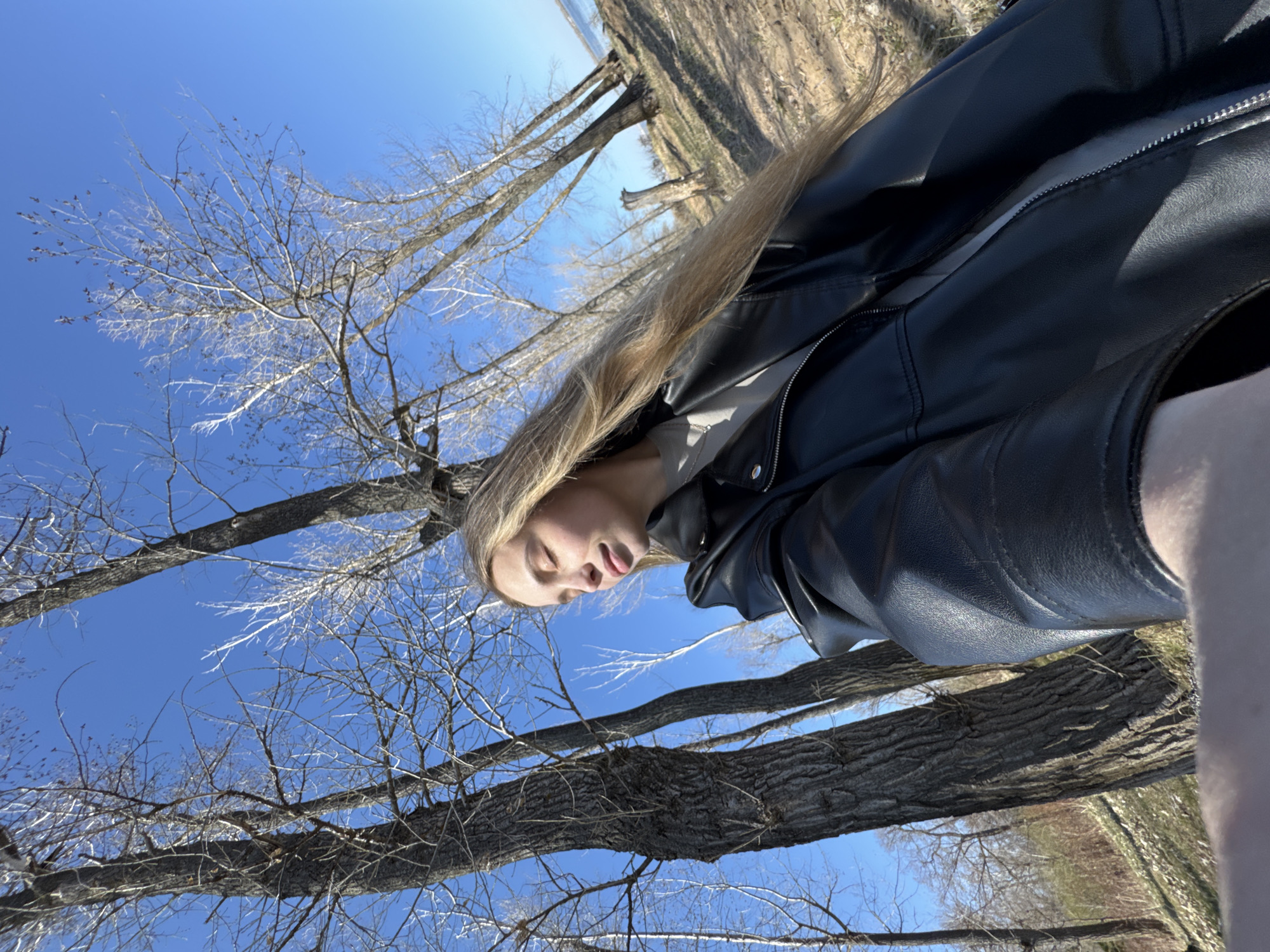 A low-angle, rotated outdoor photograph depicts a fair-skinned female wearing a black leather jacket, with long blonde hair, set against a backdrop of bare trees and a blue sky. The woman is positioned on the left of the frame, with her face appearing relaxed and eyes closed. The bare tree branches extend throughout the upper portion of the image, contrasting against the bright blue sky. The foreground features tree trunks and what appears to be a grassy ground. The location is likely Прикамский, Russia, based on the presence of trees and the geographical context.