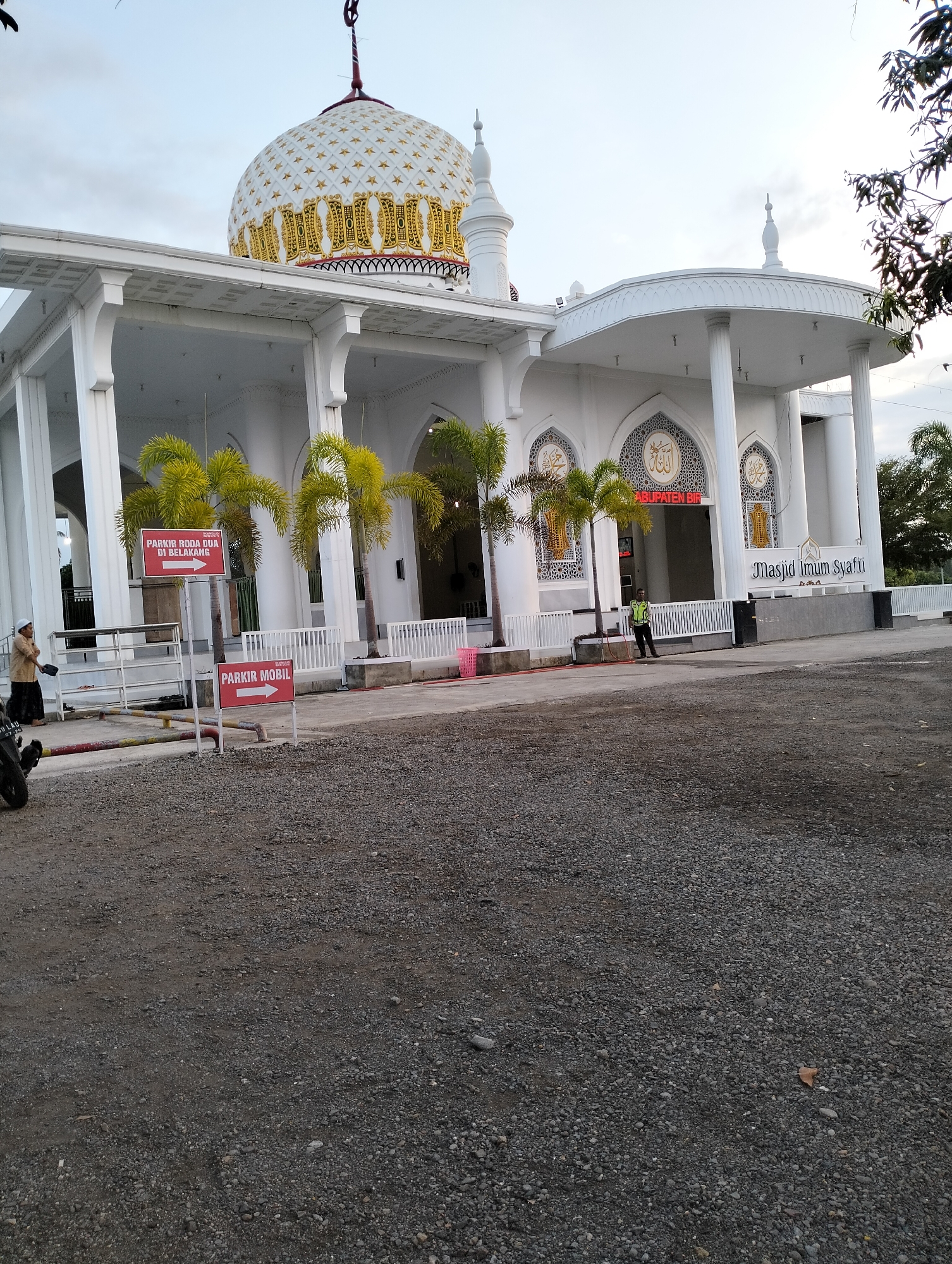 The image depicts the exterior of the Masjid Imum Syafi'i in Blangmetimu, Indonesia. The mosque is a white, modern structure with a large, ornate dome decorated with gold stars and patterns. A minaret is visible to the left of the dome. The mosque features a covered walkway with white pillars and decorative archways.
In front of the mosque, there are several palm trees. Two red signs with white text are visible: one reads "PARKIR RODA DUA DI BELAKANG" (Motorcycle parking in the back) with an arrow pointing right, and another reads "PARKIR MOBIL" (Car parking) with an arrow pointing right.
A person in a traditional long garment and head covering is seen on the left, near a motorcycle. A security guard in a high-visibility vest is standing near the entrance of the mosque on the right. The ground in the foreground is covered with gravel. The sky appears overcast.