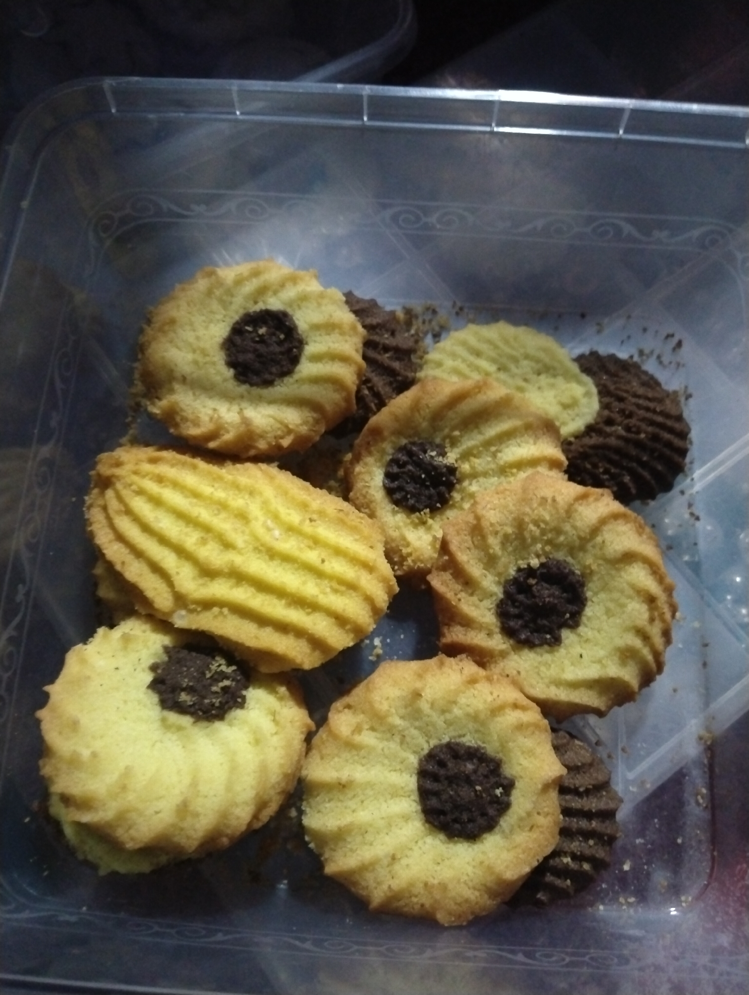 The image shows a close-up of several cookies contained within a clear plastic container, likely taken in Dera Chahal, Pakistan. The container is partially open or has a lid not fully in view, and its transparent sides reveal a subtle, decorative embossed pattern of swirls and lines.
The cookies are the primary subjects, varying in type. A majority are light yellow or golden in color, circular or oval in shape, with distinctly ridged or fluted edges. Many of these light-colored cookies feature a dark, chocolate-like circular center. Some crumbs, mostly dark brown, are scattered among the cookies and on the bottom of the container. Interspersed among these are several dark brown, almost black, cookies which also exhibit a ridged or fluted texture, suggesting a chocolate flavor or ingredient. There are approximately ten to twelve cookies visible, some stacked or partially obscured.
No people are visible in the image, nor is any specific activity taking place other than the static display of the cookies. The lighting suggests an indoor setting, but the time of day, weather, and specific location cues (beyond the container and its contents) are not discernible from the photograph. There is no visible text on the cookies or the container.