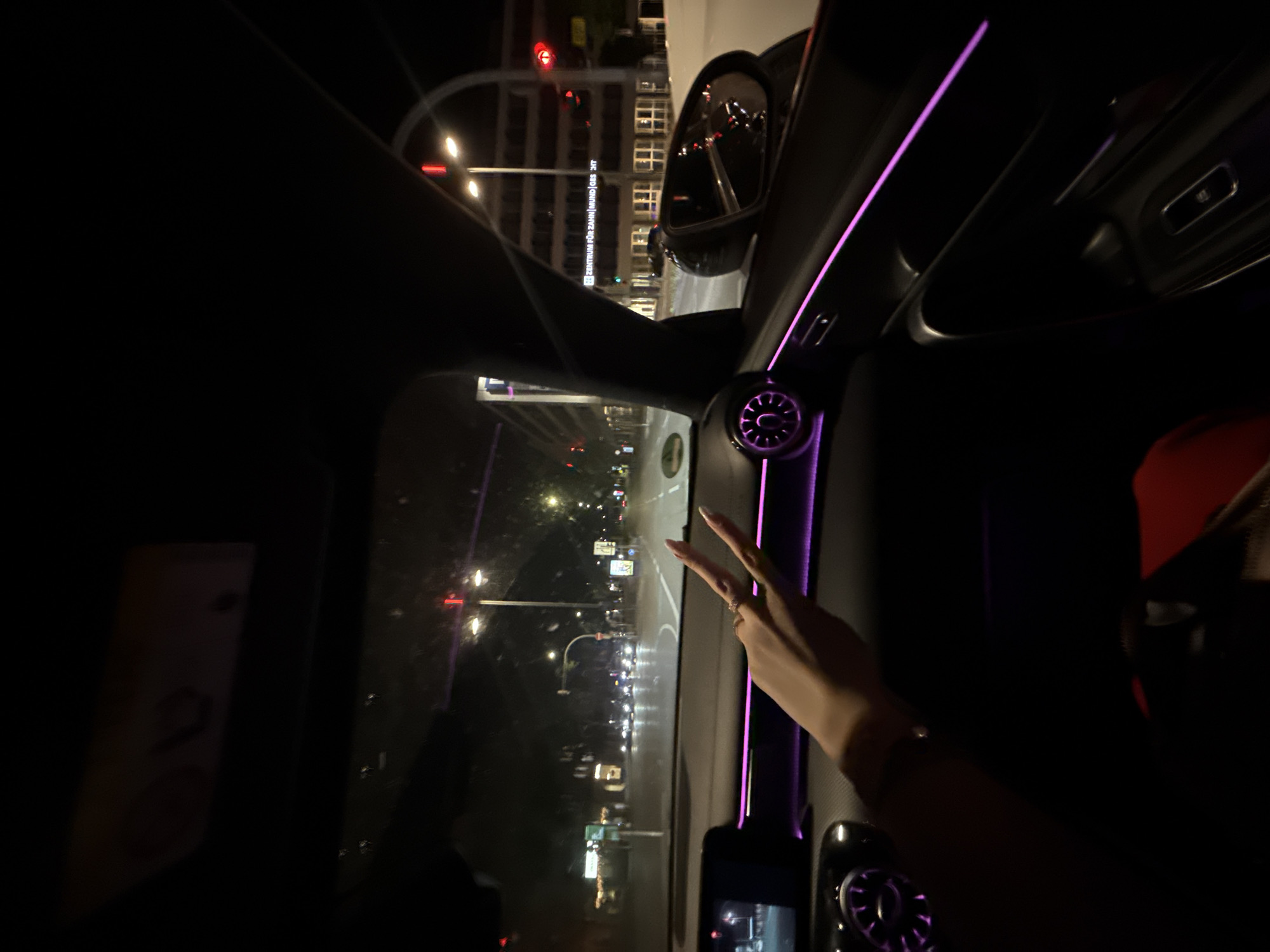 A first-person perspective shot from inside a car at night in an unknown city. The driver's hand, adorned with a ring, is visible in the foreground, making a peace sign. The car's interior features ambient purple lighting along the dashboard and door panels. Outside, city streets are visible with traffic lights, streetlights, and buildings. One building has illuminated text that reads "ZENTRUM FÜR ZAHN|MUND|GES ICHT". A side mirror reflects the street scene and car lights. The overall mood is dark with selective illumination from the city and car interior.