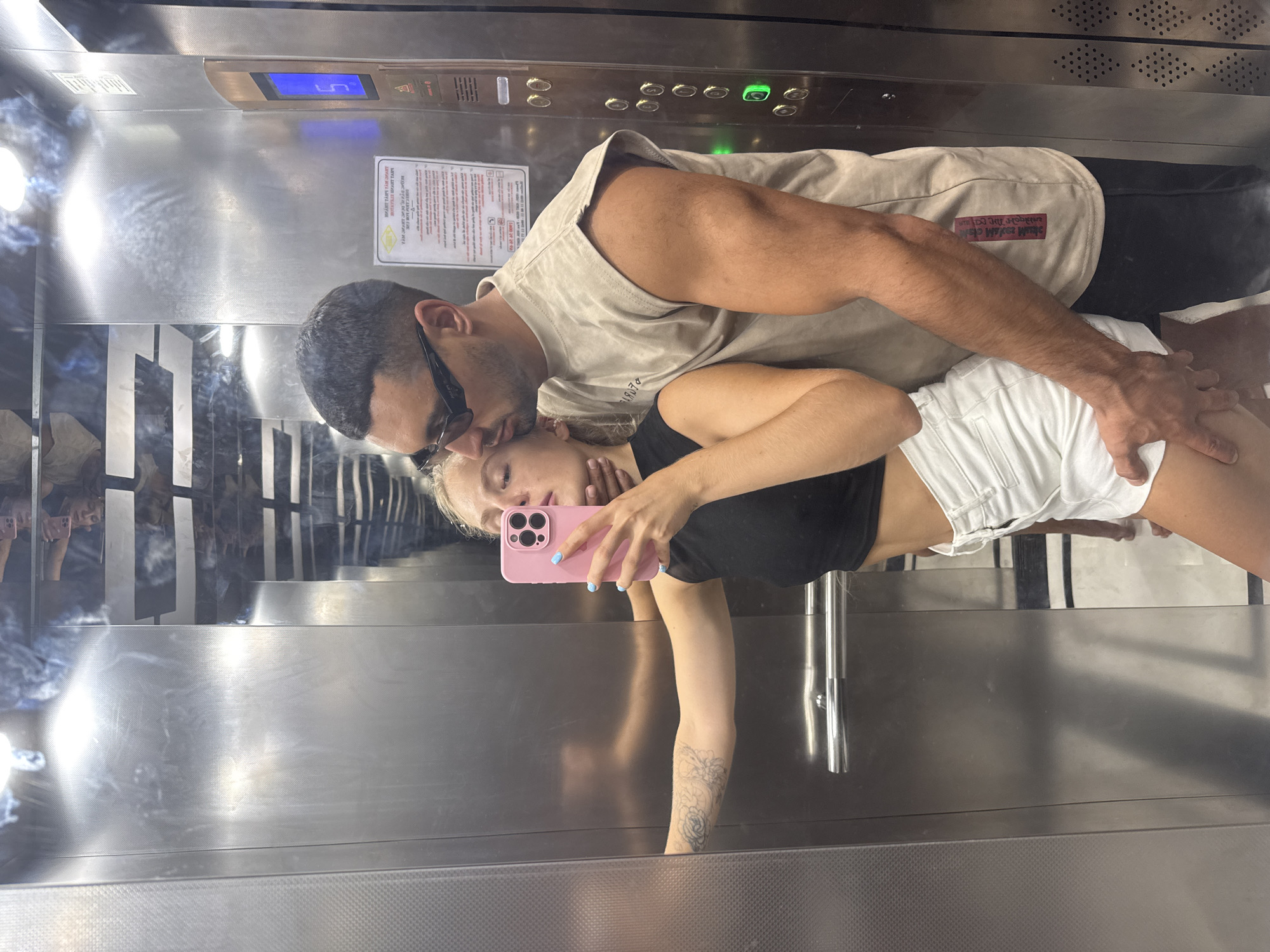 A couple is taking a selfie in an elevator. The woman is holding a pink phone with a camera in her right hand, angled to capture her and the man in the mirror. She is wearing a black crop top and white shorts. The man is wearing a beige sleeveless t-shirt and black pants. He is kissing the woman's forehead and has his arm around her. The elevator has stainless steel walls and a control panel with buttons and a digital display showing the number 5. The scene appears to be in an indoor setting, likely a building in Nha Trang, Vietnam, though the location within the city is not explicitly identifiable from the image.