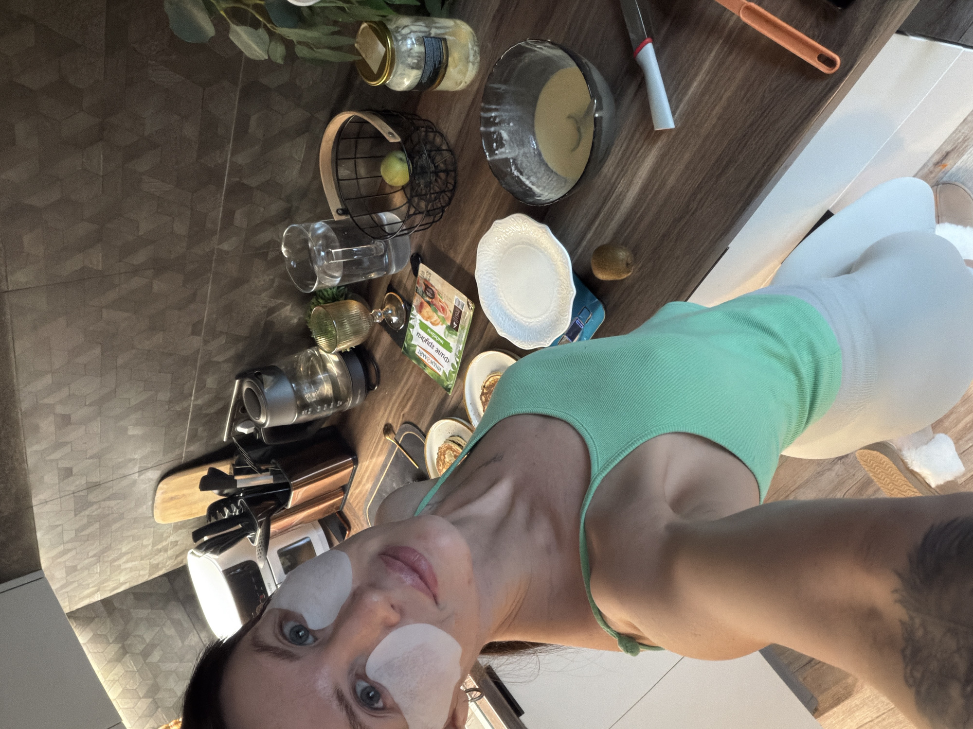 The image captures a selfie taken indoors, seemingly in a kitchen, located in Perm, Russia. A woman with light skin, sporting eye patches, is in the foreground, wearing a light green tank top and white leggings. Her left arm features a tattoo.
The countertop is a dark wood-grain style. Various kitchen items are scattered across it including: a glass jar, a black wire basket with a green apple inside, a glass cup, a potted succulent, a silver kettle, a knife block with utensils, a white mixer, a white plate, an open package of "Фит Зарянка" tea, a kiwi, a light blue box, and two small plates with pancakes. A ceramic bowl contains a light-colored mixture, possibly batter.
The kitchen also includes a plant with green foliage and an orange spatula near the bowl. The floor is tiled with a hexagonal pattern. A white kitchen cabinet is visible in the background.