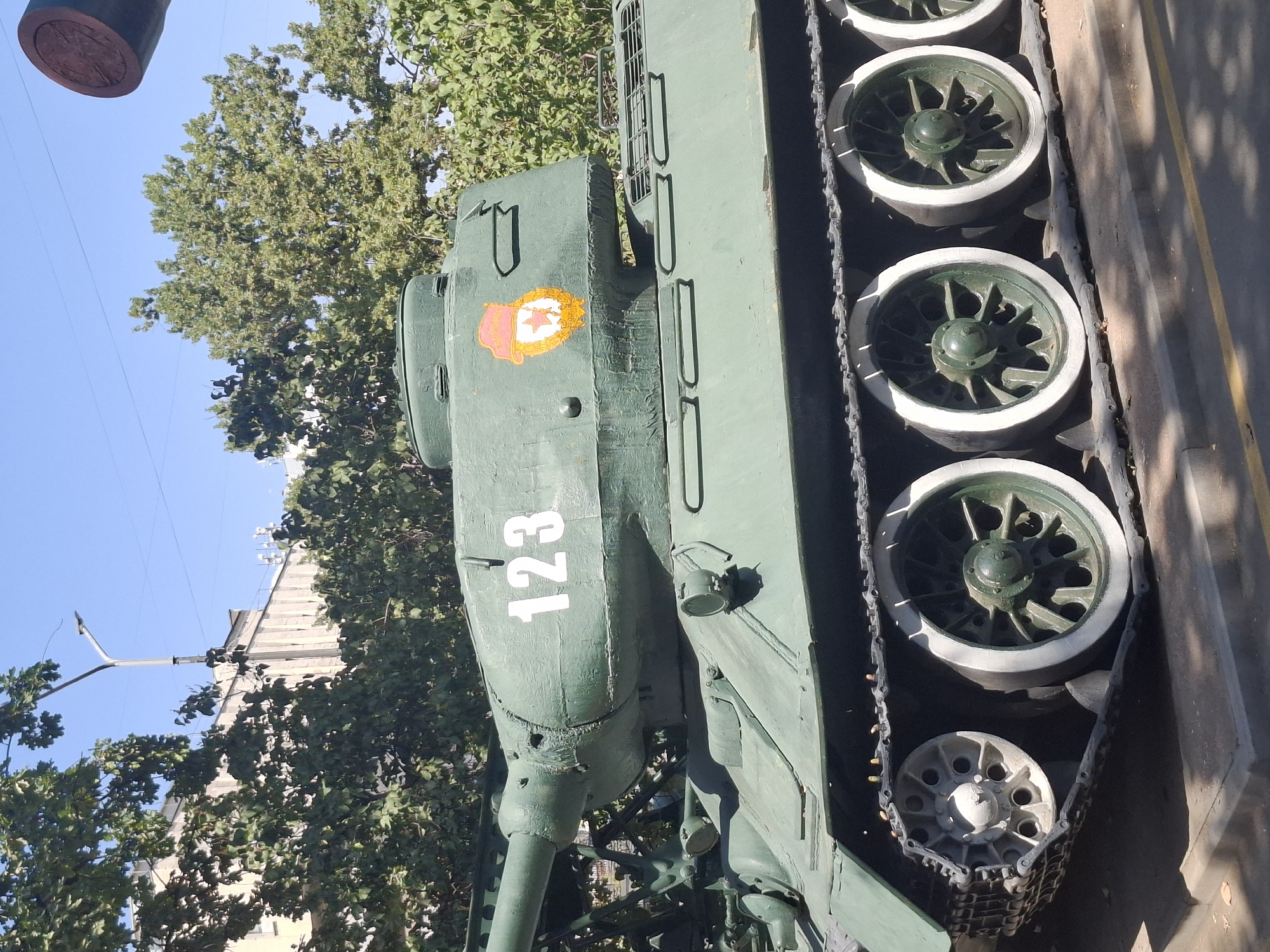 A green Soviet-era T-34 tank is on display outdoors in Almaty, Kazakhstan, situated at a monument. The tank is painted a matte green color, with the number "123" painted in white on its turret. A circular emblem, depicting a red star within a wreath and a stylized hat, is also visible on the turret. The tank's tracks and road wheels, painted green with white rims, are prominent. The background includes lush green trees, a building with a light-colored facade, and a clear blue sky. The tank is positioned on a concrete base, with a yellow line marking the edge of a paved area. The lighting suggests it is daytime, with shadows cast on the ground. No people are visible in the frame.