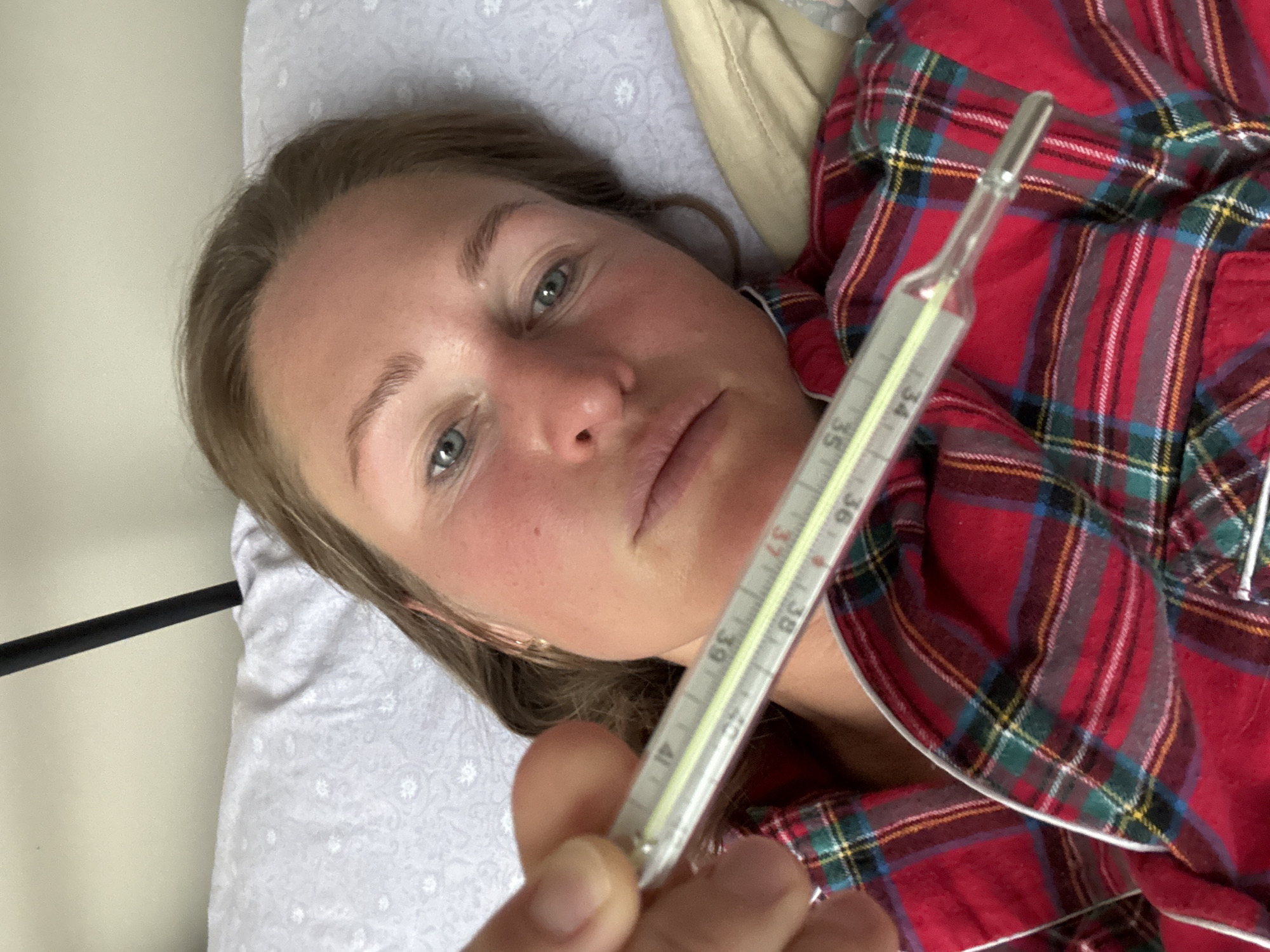 A woman is lying in bed, covered in a red and green plaid pajama top. She is holding a mercury thermometer up to the camera. The mercury in the thermometer reads approximately 38.5 degrees Celsius. Her face appears flushed. She is lying on a white pillow with a light grey patterned pillowcase. The image was taken in an indoor environment that may be located in the city of Chaikovsky, Russia.