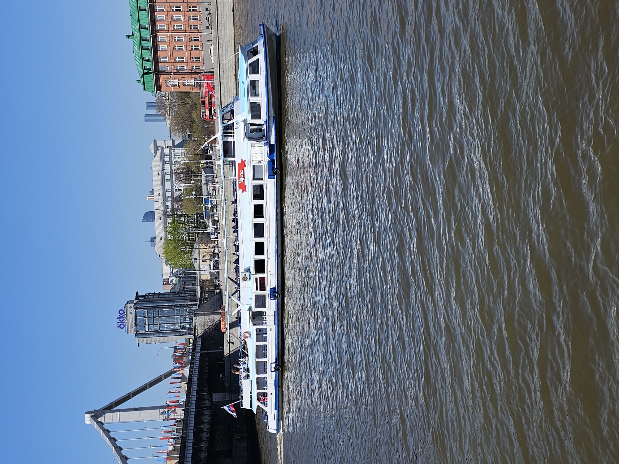 Here is a description of the image:
The image depicts a river scene in Moscow, Russia. A white and blue passenger boat is docked alongside a concrete embankment. The river is a muddy brown color with textured ripples. In the background, a cityscape emerges, featuring buildings of varying sizes and architectural styles. The cityscape includes modern glass structures, red brick buildings with green roofs, and other nondescript buildings. In the foreground, a bridge adorned with colorful flags spans the river. The sky is a clear, bright blue.