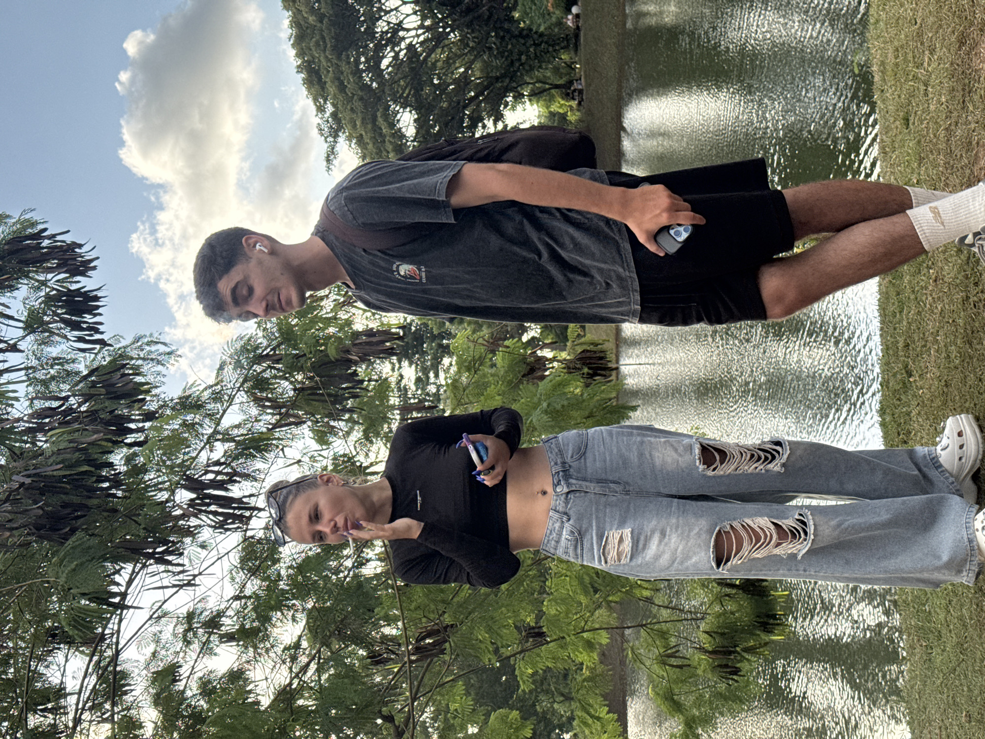 Two people, a man and a woman, are standing in a park-like setting near a body of water. The woman, positioned closer to the viewer, is wearing a black long-sleeved crop top and ripped blue jeans. She is holding an object in her left hand and appears to be smoking. The man is wearing a dark grey t-shirt and black shorts. He is holding a phone in his right hand and has an earpiece in his left ear. The background features trees, grass, and a body of water. The sky is visible with some clouds. The city of Kuta Selatan, Indonesia is not clearly identifiable in the image but is mentioned as a possible location.