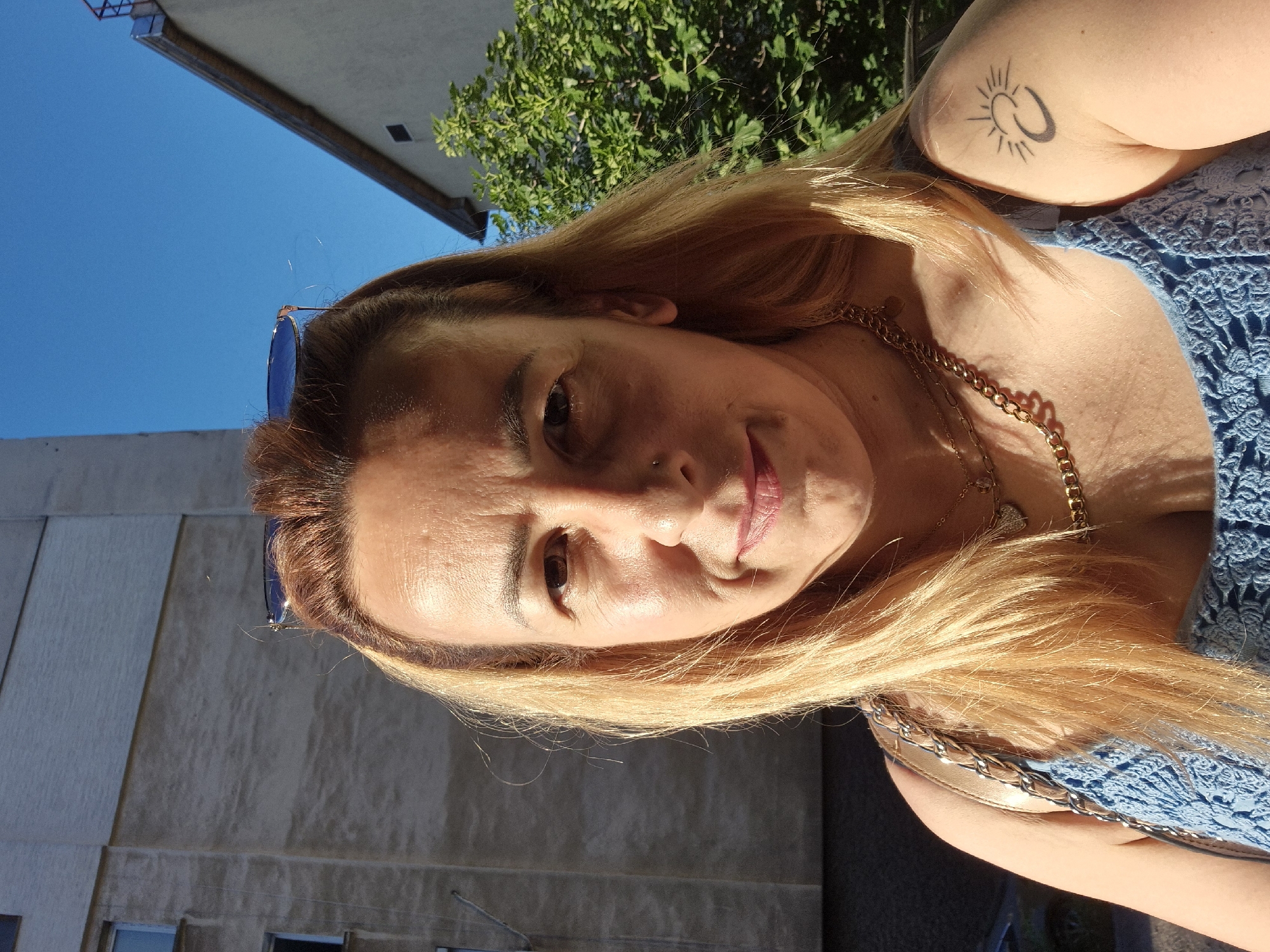 A frontal, medium shot shows a woman in her late 40s with light brown hair, fair skin, and light brown eyes. She is wearing a blue crocheted top and gold necklaces, one with a heart-shaped pendant. A silver chain strap from a purse is visible on her left shoulder. She has a tattoo of a crescent moon and sun on her right upper arm. Sunglasses are perched on her head. The woman is smiling slightly and looking at the camera. Behind her, a building with a light-colored facade and windows is visible, along with a bright blue sky and green foliage. The lighting suggests it is a sunny day, with harsh shadows cast on the building and the woman's face.