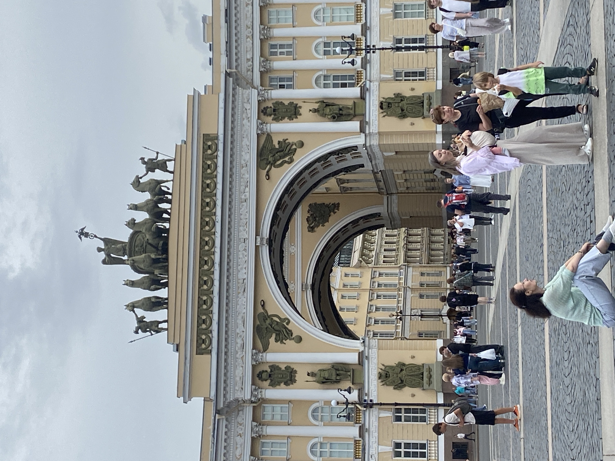 The image depicts the General Staff Building in Palace Square, Saint Petersburg, Russia, under an overcast sky. The iconic triumphal arch of the building is a prominent feature, adorned with classical sculptures and architectural details. A chariot with horses and a winged goddess, symbolizing glory, crowns the arch.
In the foreground, a diverse group of people are visible. To the right, a woman with glasses is sitting on a low ledge, looking to the side. To her left, a woman in a light pink shirt and wide-leg beige pants holds a baby in a wrap and is in conversation with another woman holding a paper bag. A young boy in a green and white shirt stands in front of them, also holding a snack. In the middle ground, other pedestrians are walking across the cobblestone square, some carrying bags and backpacks. The architecture of the surrounding buildings is visible through the archway, showcasing a classic European urban landscape. The overall atmosphere suggests a public gathering or a tourist area.