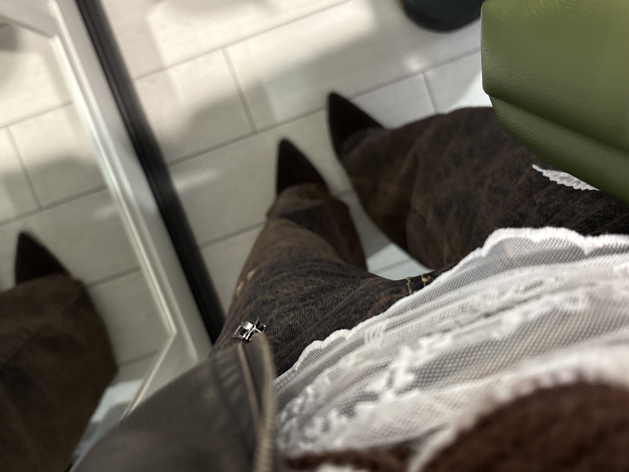The image shows a person standing on a white tiled floor, presumably indoors. The person is wearing brown jeans and pointy brown shoes. A light green bag is visible near the person's leg. Part of a white lace garment and a grey zipper are also present. A mirror with a white frame is on the left. The location is not specified.