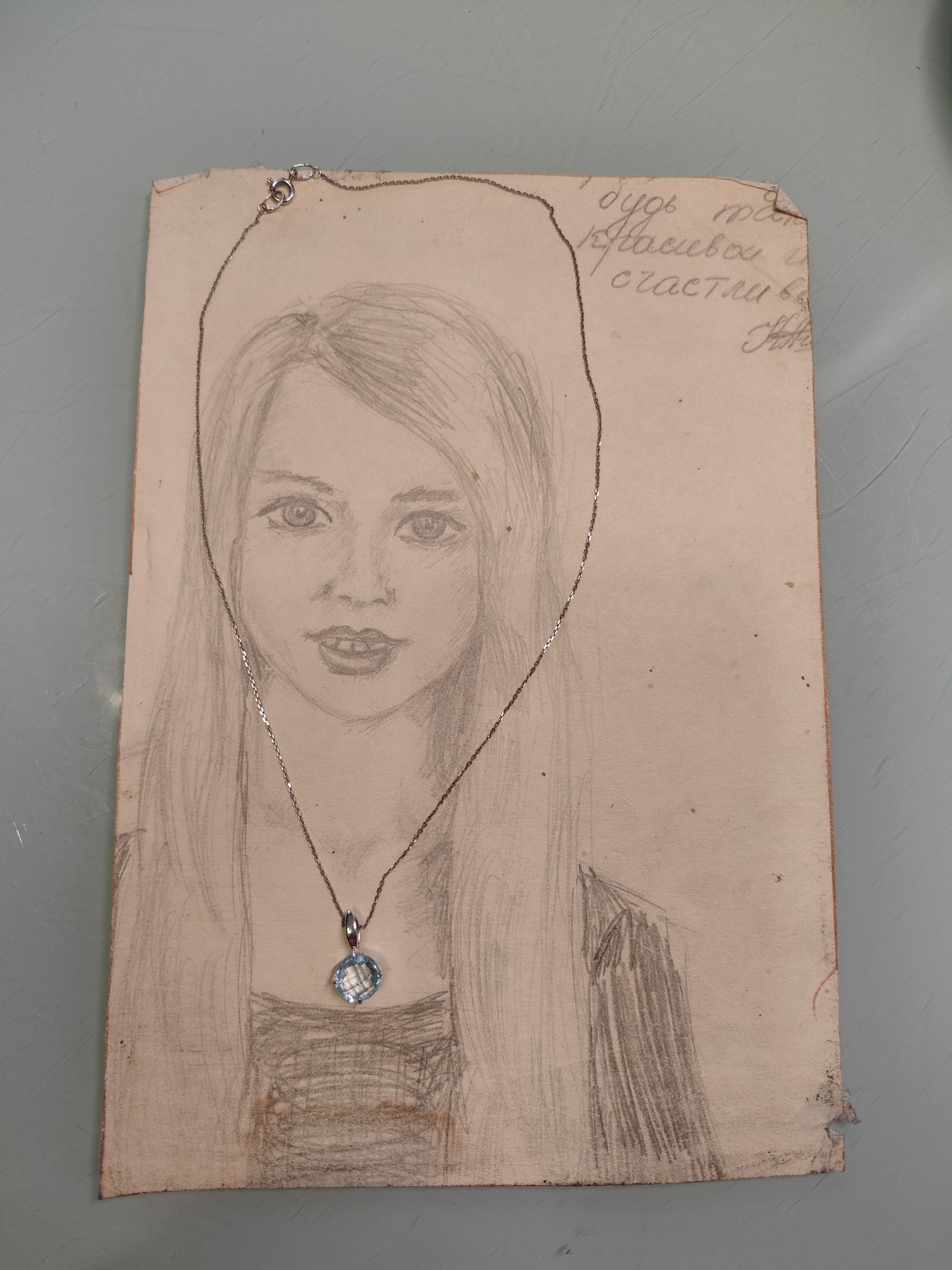 A pencil sketch of a young woman's face is displayed on a piece of aged paper. The sketch depicts her with long, flowing hair and a slightly open mouth. A delicate silver chain necklace with a square, light blue gemstone pendant is draped over the paper, positioned in front of the sketched face. To the upper right of the sketch, handwritten text in Cyrillic script is visible. The paper is worn and slightly torn at the edges. The background is a plain, muted gray surface. The scene is not identifiable as being located in Gvardeysk, Russia.
