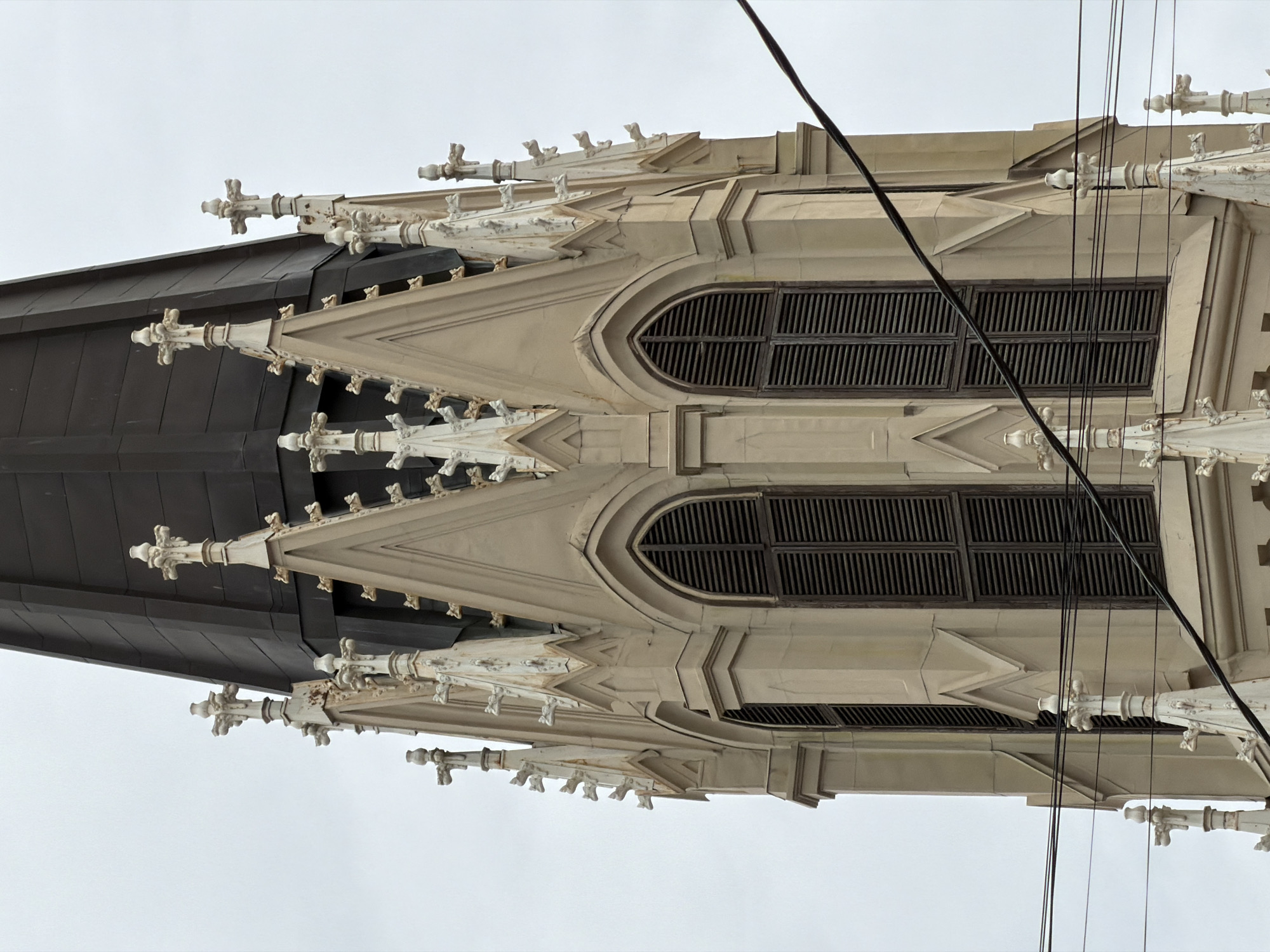 The image shows a detail of the Saint Isaac's Cathedral in Saint Petersburg, Russia. The view is an upward angle of the cathedral's architecture, highlighting the gray roof, light beige stone facade, windows, and decorative elements, along with powerlines. The weather appears overcast.