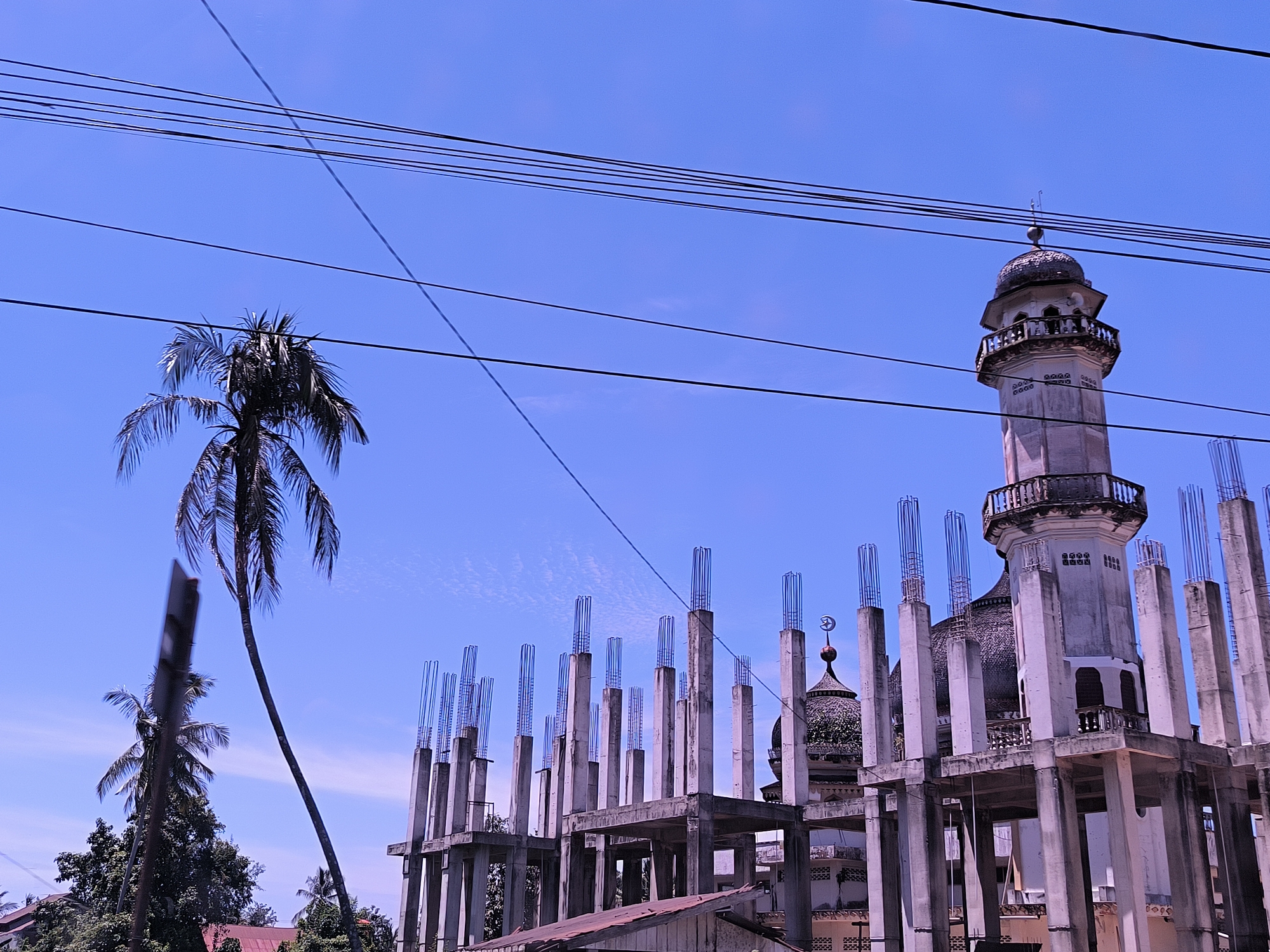The image shows a building under construction in Keudealue Ie Puteh, Indonesia. The structure appears to be an expansion or renovation of an existing mosque. The concrete framework of the new construction surrounds the older, completed sections of the mosque, including a minaret and a dome. Reinforced steel bars protrude from the tops of the concrete columns. Overhead, a network of power lines crisscrosses the sky. A tall palm tree stands to the left of the building. Another shorter palm tree is next to the structure. The sky is blue and clear.