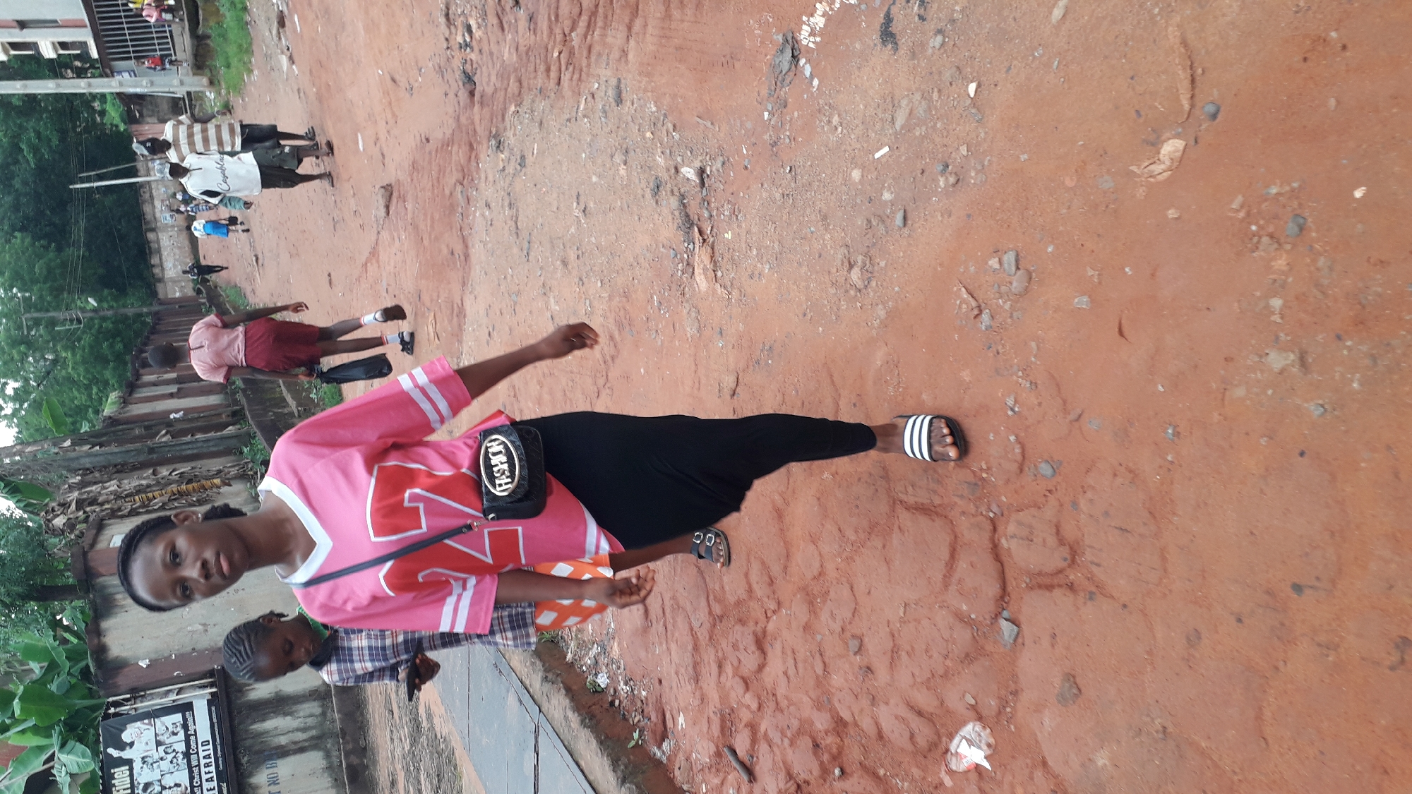The image depicts a young woman standing on a red, uneven dirt road. She is wearing a pink and white oversized jersey with the number 22, black leggings, and black and white striped sandals. A small black purse labeled "FASHION" is slung across her body. Behind her is another child, partially visible, wearing a plaid shirt. In the background, several other people can be seen walking on the same road. There is a building with peeling paint and posters on its side to the left. The environment suggests an urban setting in the city of Onitsha, Nigeria due to the red soil and infrastructure.