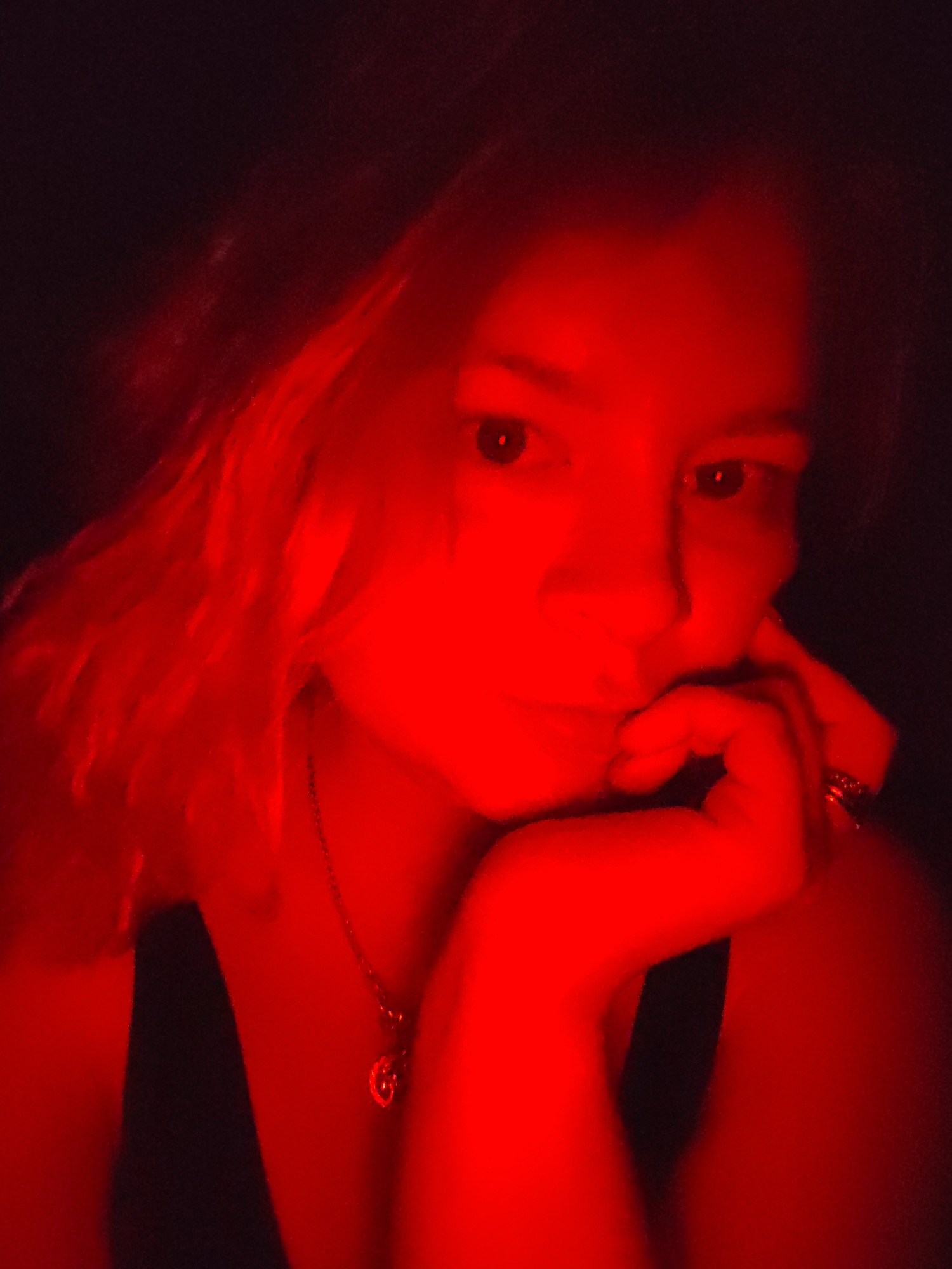A close-up shot of a woman with fair skin and reddish-blonde hair illuminated by a strong red light source. She is resting her chin on her hand, which has a ring visible on one of her fingers. She wears a dark-colored top and a necklace with a pendant. The background is dark, obscuring details. The image offers no information regarding the city of Izhevsk, Russia.