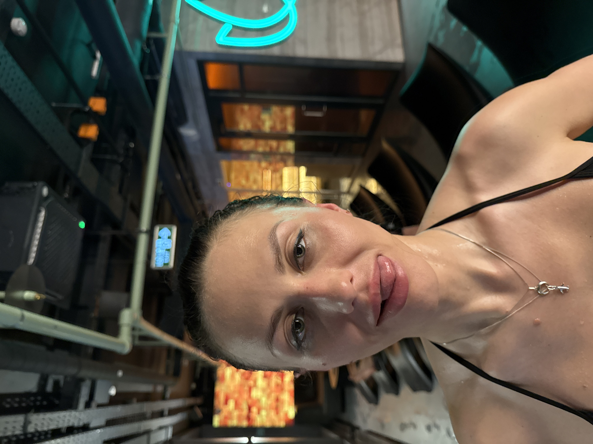 A woman is positioned in the foreground of an interior space, likely captured from a low angle, with a slight tilt to the camera's orientation. She is wearing a black top with thin straps and a delicate silver necklace. Her face is visible, exhibiting a light sheen suggesting moisture or sweat.
In the background, the setting appears to be an industrial or commercial space, indicated by metal structures and pipes. There is an interplay of colors, with a bright orange or amber glow coming from an unseen source, possibly lighting, and a contrasting blue hue from neon lights overhead. The structural elements and lighting suggest an urban environment. Based on the overall composition and provided context, this location could potentially be within the city of Saint Petersburg, Russia.