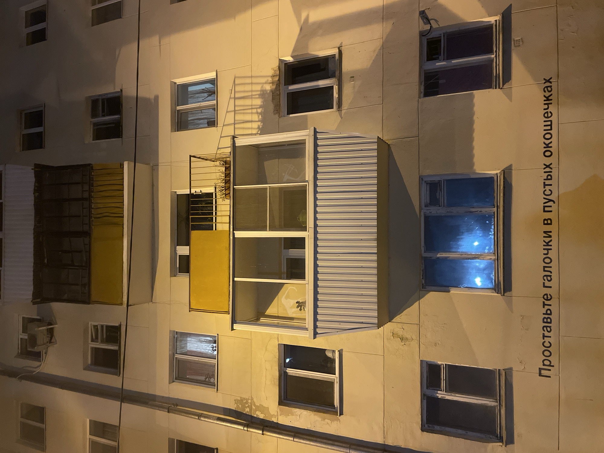 The image displays a low-angle, nighttime view of a multi-story residential building facade in Yekaterinburg, Russia. The exterior is a pale yellow or beige, exhibiting weathered patches and peeling paint under artificial yellowish illumination, which casts distinct shadows.
The facade features numerous rectangular windows across multiple floors. Some windows appear dark, while others reveal faint interior lights or reflections. On the upper-left, a wall-mounted air conditioning unit is visible. A vertical metal downspout runs along the left side of the building.
Two distinct balconies are present:
1. An open-air balcony on an upper floor, characterized by a dark metal railing and a yellow-painted lower panel.
2. A large, enclosed, glass-paneled loggia on a lower floor, featuring a white, vertically ribbed lower section. Above this loggia, a smaller open-air balcony with a black metal railing and a visible planter box with plants is situated.
At the lowest visible level of the building, two rectangular windows are prominent. The left window is dark, while the right window emits a bright, distinct blue light from within. Below these windows, black Cyrillic text is painted directly onto the facade, stating: "Проставьте галочки в пустых окошечках" (Prostav'te galochki v pustykh okoshechkakh), which translates to "Put checkmarks in the empty boxes." No individuals or overt actions are depicted.