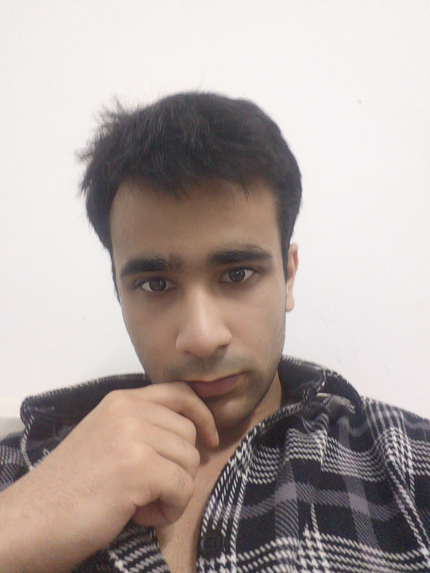 A head-and-shoulders portrait of a young adult male with dark hair. He is wearing a black and white plaid shirt and his hand is touching his chin. The background is a plain white wall. The location in the city of Chongqing High-tech Zone, China is not discernable from this image.