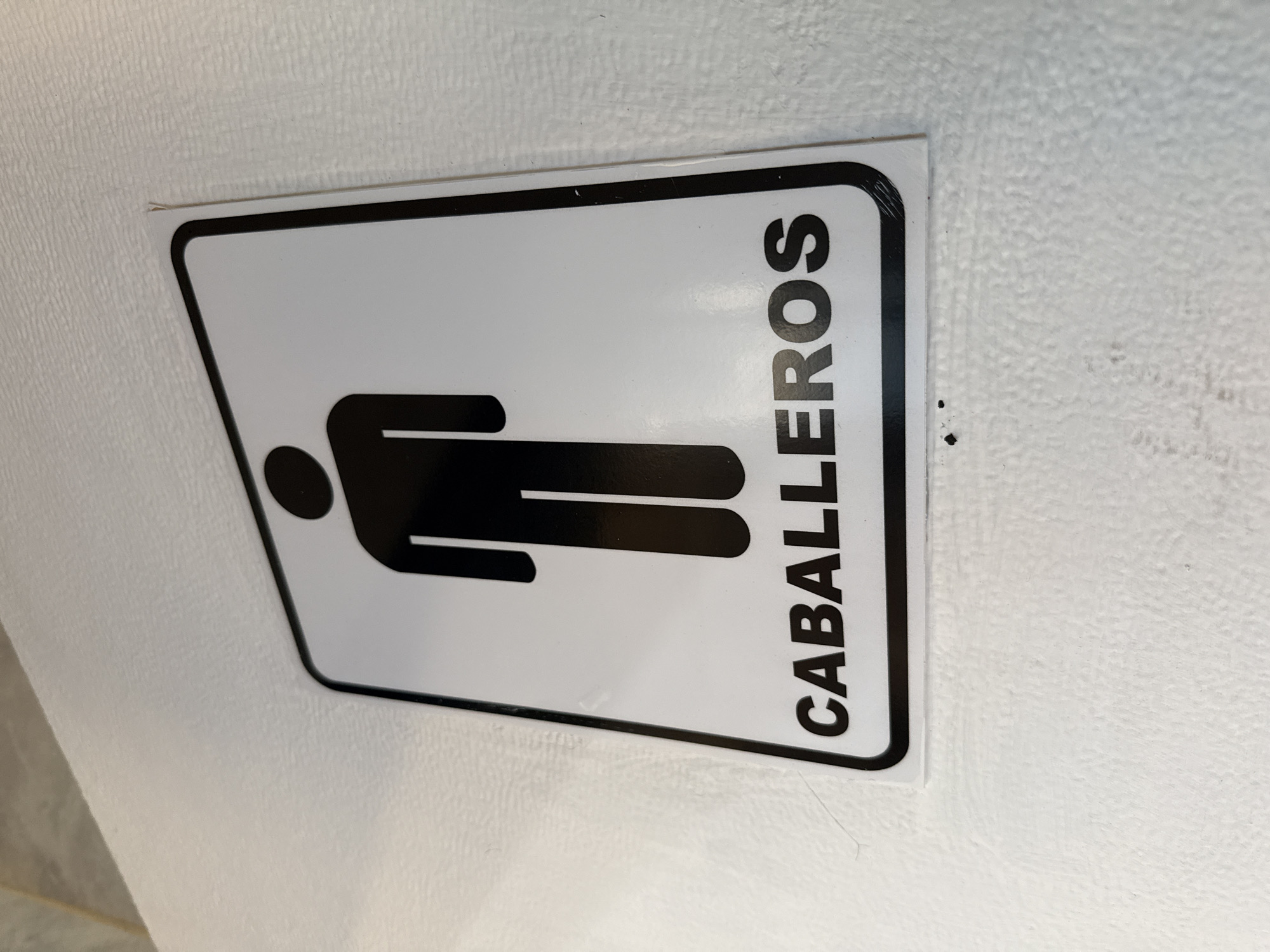 A sign for the men's restroom (Caballeros in Spanish) is affixed to a white wall. The sign is white with a black border and black text. A stylized figure of a man is displayed on the left. The location is Tinaquillo, Venezuela.