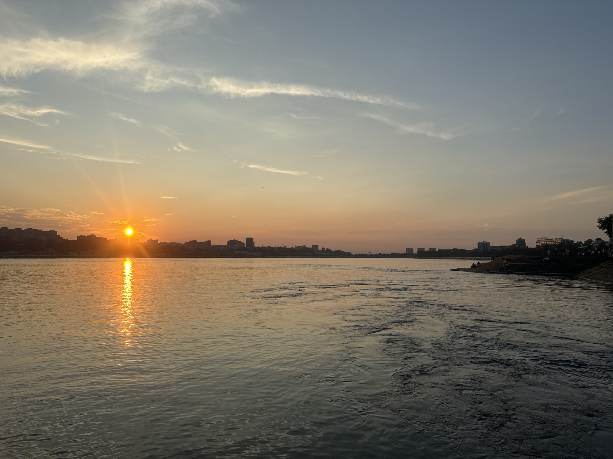This is a wide-angle photograph of the Angara River in Irkutsk, Russia, taken during sunset. The sun, a bright orange orb, is low on the horizon, casting a shimmering reflection across the water. The sky is a gradient of soft blues and oranges, with wispy clouds stretched across it. On the far bank of the river, a cityscape is visible, silhouetted against the fading light. Buildings of varying heights line the river, suggesting an urban environment. The water itself is dynamic, with ripples and currents creating patterns on its surface. Towards the right of the frame, a sloped embankment with tiered steps is visible, with several figures seated or walking on it. A tree stands at the far right edge of the embankment. The scene captures a tranquil moment as day transitions into evening.