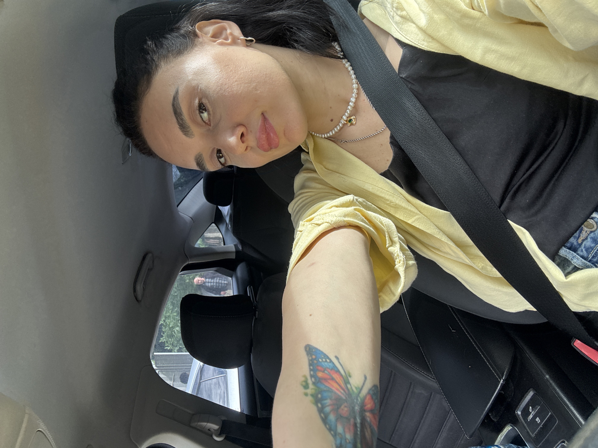 A woman sits in the driver's seat of a vehicle, fastened with a black seatbelt. She is wearing a black top under a yellow shirt, a pearl necklace, and has a butterfly tattoo on her arm. She is looking at the camera. The car's interior is visible, along with a glimpse of the outdoors through the windows. The photo does not contain enough information to determine the location in the city of Tbilisi, Georgia.