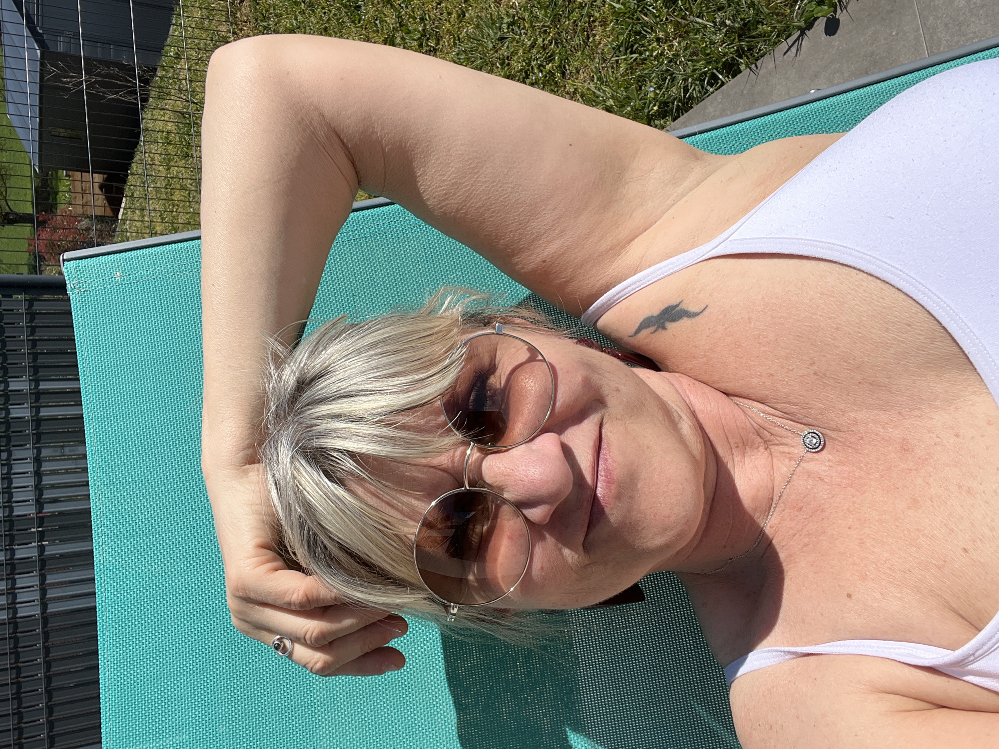 A woman wearing a white tank top and round, brown-tinted sunglasses is reclining on a turquoise-colored lounge chair outdoors. She has blonde hair with darker roots and is wearing a ring on her finger and a necklace. A small tattoo is visible on her upper chest. The scene appears to be in a backyard with grass and a black metal fence. The location is Chambœuf, France.
