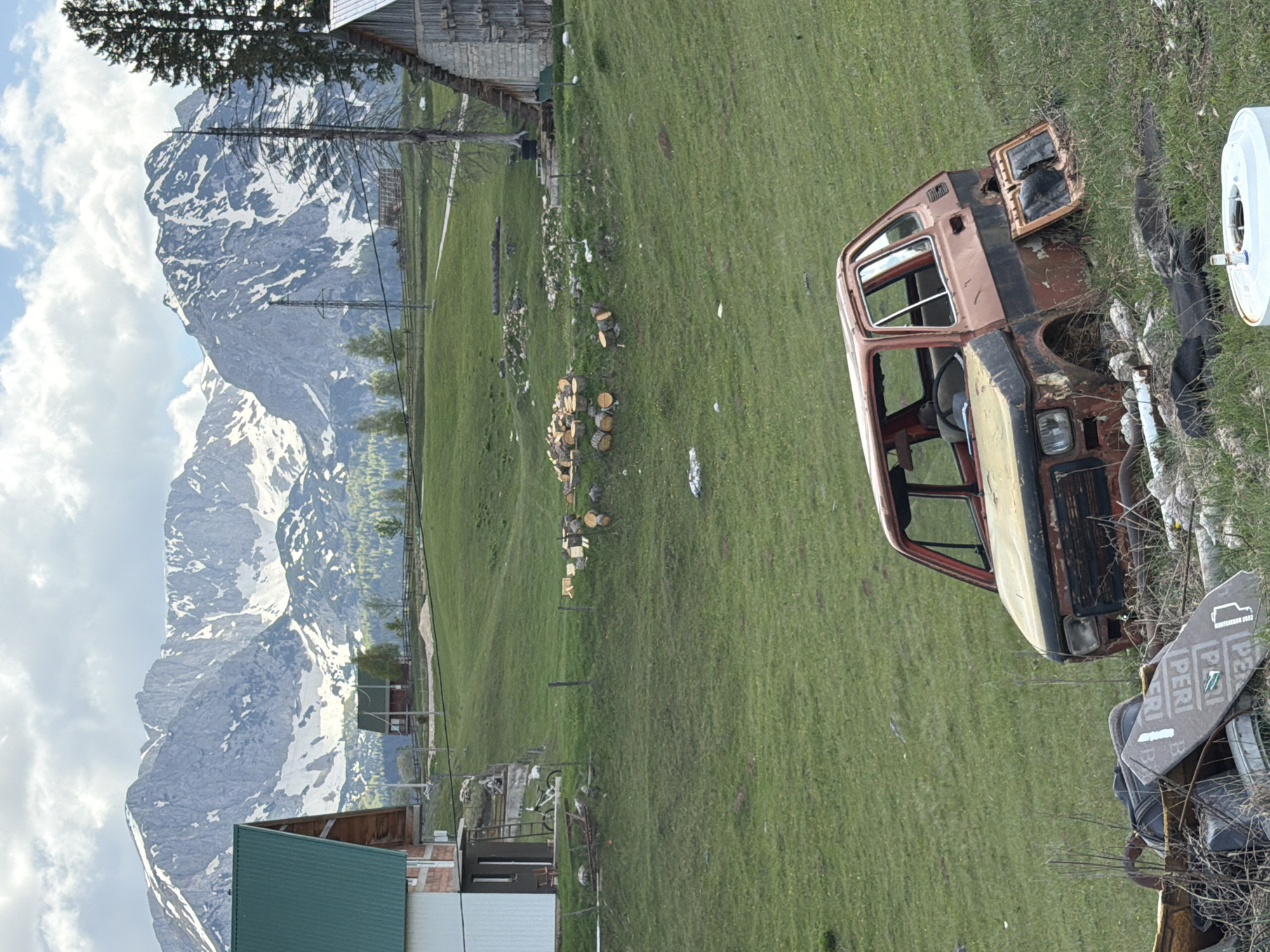 The image shows a rural scene in Žabljak, Montenegro, with a backdrop of mountains with snow. In the foreground is the rusted cabin of a vehicle on a grassy field. To the left, there is a wooden building and chopped firewood. A gray surface with white lettering is located next to the car. The weather appears to be clear, with a partly cloudy sky.