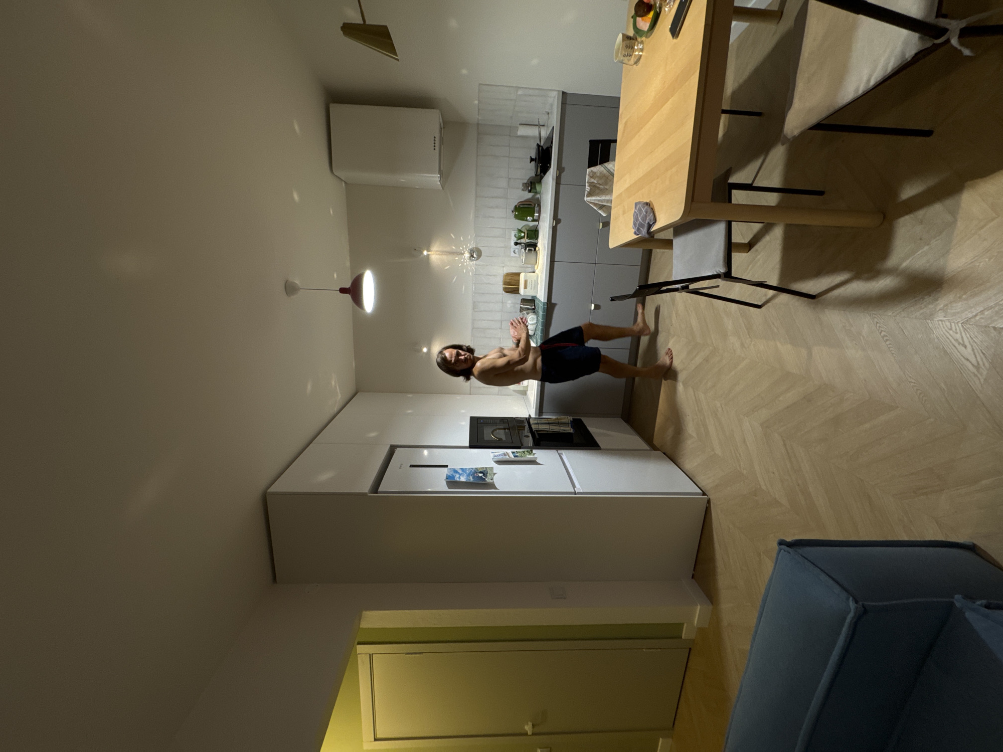 The image captures a modern apartment interior, likely located in Izhevsk, Russia, encompassing a kitchen, dining area, and a glimpse of a living space. The room is brightly illuminated by artificial lighting, creating a clean and contemporary ambiance. The floor features a light wooden herringbone pattern, and the walls are a neutral, light color.
In the mid-ground, a man with long dark hair, shirtless and wearing dark shorts, is performing an athletic pose resembling a push-up or plank, with his hands together on the floor and his body elevated. He looks directly at the camera with a composed expression, indicating a deliberate pose.
The kitchen area to his left is equipped with grey lower cabinets, white upper cabinets, a white countertop, and a tiled white backsplash. Visible appliances include a white range hood, a black stovetop, a microwave, and a white refrigerator adorned with magnets, one clearly depicting a cow in a field. Several distinctive green kitchen appliances are lined up on the counter. Overhead, a red-accented pendant light and a reflective, disco ball-like fixture illuminate the space.
To the man's right, a light wooden dining table is set with a bowl of fruit, glasses, a smartphone, and a cloth. Two chairs with light cushions and dark frames are placed around the table. A section of a dark blue upholstered armchair or sofa is visible in the foreground. In the background, a doorway leading to a room with light green walls can be seen. The overall scene suggests a well-maintained and stylish urban dwelling.