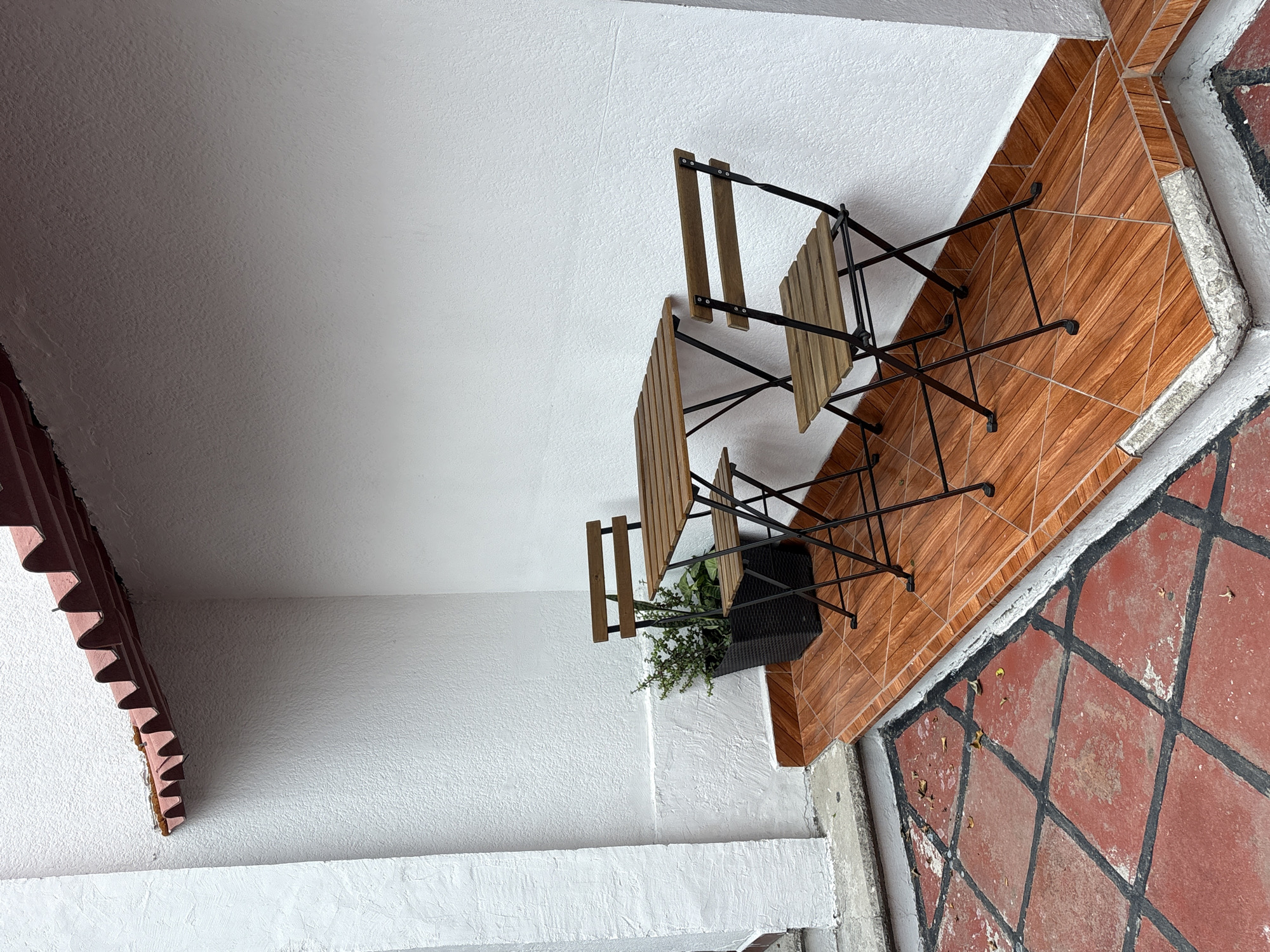 This is an indoor scene, possibly a patio or balcony, in Puebla, Mexico. Two small, wooden bistro-style chairs with black metal frames are positioned on reddish-brown tiled flooring. A potted plant sits between the chairs. The walls are textured white, and the ceiling is also white. The edge of a red tiled roof is visible in the upper left corner of the image. The scene is well-lit. Additional red-and-black tiles are visible at the bottom of the image.