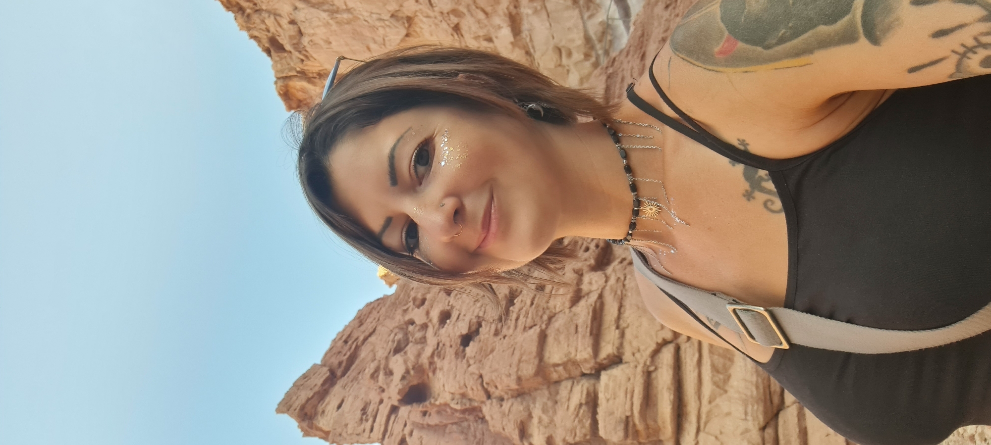 A selfie captures a woman in the city of Hevel Eilot Regional Council, Israel. She has fair skin, brown hair, and is wearing glitter on her face. Her left shoulder is visible, revealing a tattoo, and she is wearing a black tank top and a necklace with silver chains. A strap is across her chest. The background shows a blue sky and a large rock formation.