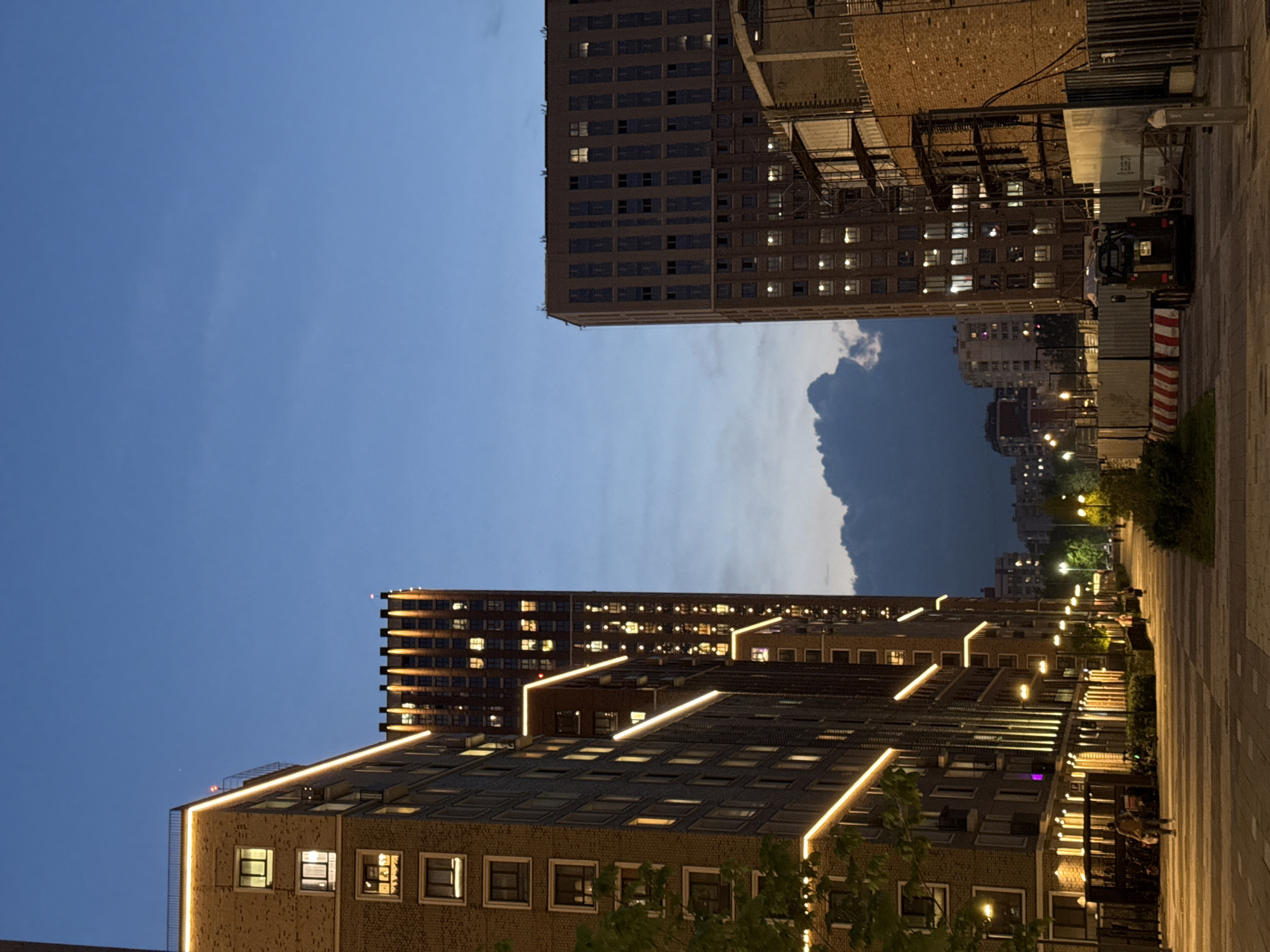 The image depicts a view in the city of Khimki, Russia, at dusk or twilight. Two tall buildings, constructed with brick facades and featuring numerous windows with illuminated interiors, frame a narrow view of the sky. The buildings are additionally embellished with light fixtures along their edges. A partially visible pathway, constructed with patterned pavers, extends alongside one building, marked with streetlights and greenery. In the distance, additional buildings are dimly illuminated. A large, dark cloud formation occupies the sky between the buildings. Construction equipment and temporary barriers are seen near the upper building. The image appears to be taken from a low vantage point, emphasizing the height of the buildings.