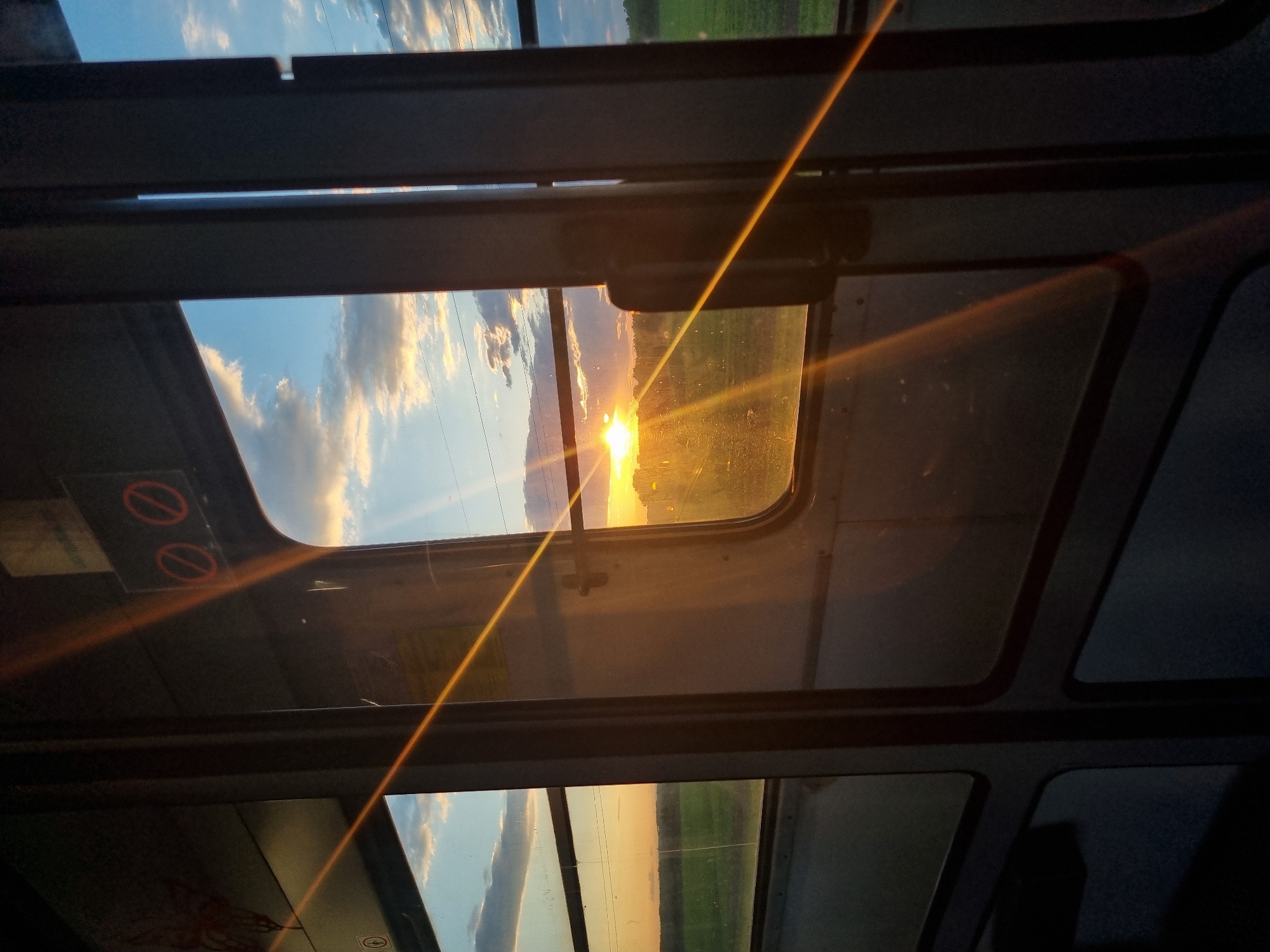The image depicts a view through a paneled window, likely a train or bus, with the sun setting. The scene outside the window shows a landscape with green fields under a sky with clouds. The sun is positioned near the horizon, creating a bright glare and causing reflections on the window surfaces. In the foreground, the window frame consists of dark, rectangular panels with rounded corners. There are also warning signs on the interior glass, which are blurred by reflection. The city, specific location, and interactions are unknown.